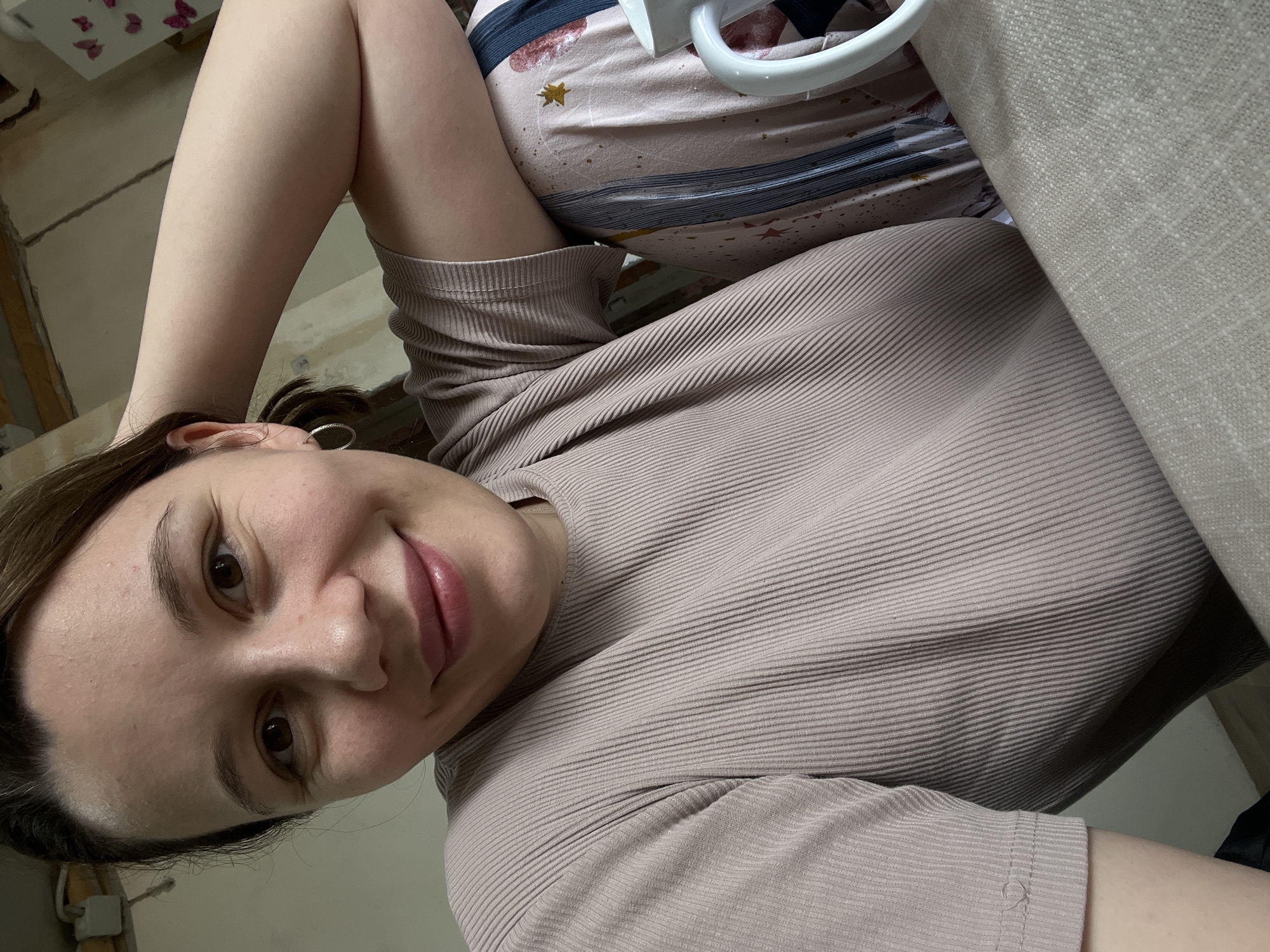 A woman with brown hair, wearing a light brown ribbed shirt, lies down and looks into the camera. She is on a surface covered with beige linen, next to pajamas with a star and cloud pattern and a white plastic object. The location is possibly a room with partially finished walls. No information is available to verify the city and country mentioned.