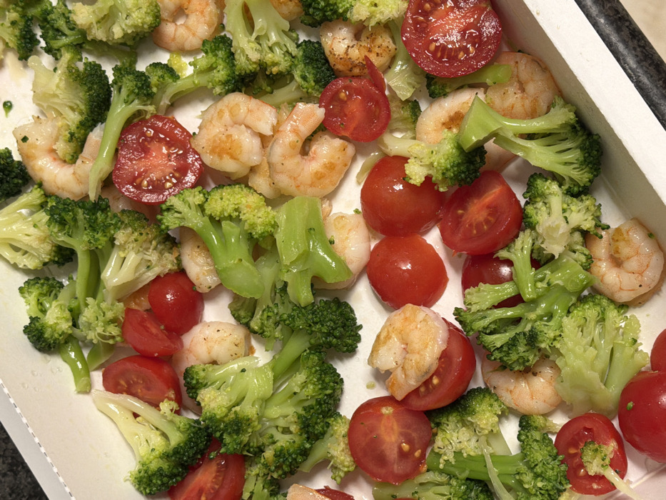 The image displays a close-up, top-down view of a colorful meal or dish, presented in a white, rectangular container or tray with slightly raised edges. The primary ingredients are cooked shrimp, broccoli florets, and cherry tomatoes, mixed and scattered throughout the container.
The shrimp are a pale pinkish-orange color, curled, and appear to be fully cooked. Some have visible flecks of seasoning, possibly pepper or herbs. They vary in size but are generally medium-sized.
The broccoli consists of bright green florets and some stem pieces. The broccoli appears to be cooked, possibly steamed or lightly roasted, maintaining its vibrant color and structure.
The cherry tomatoes are a deep, bright red. Many are cut in half, revealing their juicy interior and small seeds, while others are whole and round. They are interspersed evenly among the shrimp and broccoli.
The combination of ingredients suggests a healthy and light meal, possibly a stir-fry, salad component, or roasted dish. The food is arranged loosely, not neatly plated, implying it might be served family-style or as a prepped meal.
The white container provides a clean contrast to the vibrant colors of the food. A dark, possibly stone or countertop surface, is visible in the background along the bottom right and left edges of the tray. The lighting is bright and even, illuminating the food clearly. No text is visible in the image.
