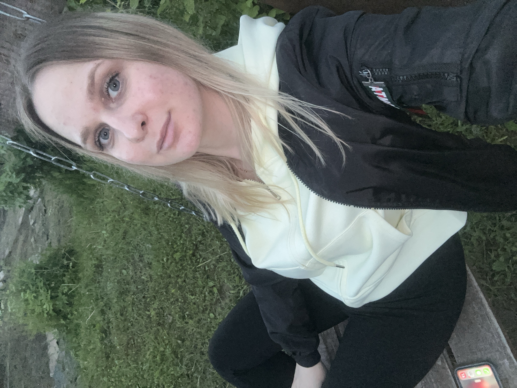 A young adult female with blonde hair and blue eyes is seated outdoors in the city of Chaikovsky, Russia, looking directly at the camera. She is wearing a light yellow hooded sweatshirt under a black bomber jacket, which features a zippered pocket on the left arm with a visible "ATW" tag. She is also wearing black pants. A mobile phone displaying app icons rests on a wooden surface to her left. Metal chains are visible on either side of her head, consistent with being on a swing. The background consists of green grass and sparse foliage, indicating a natural, outdoor setting under dim lighting.