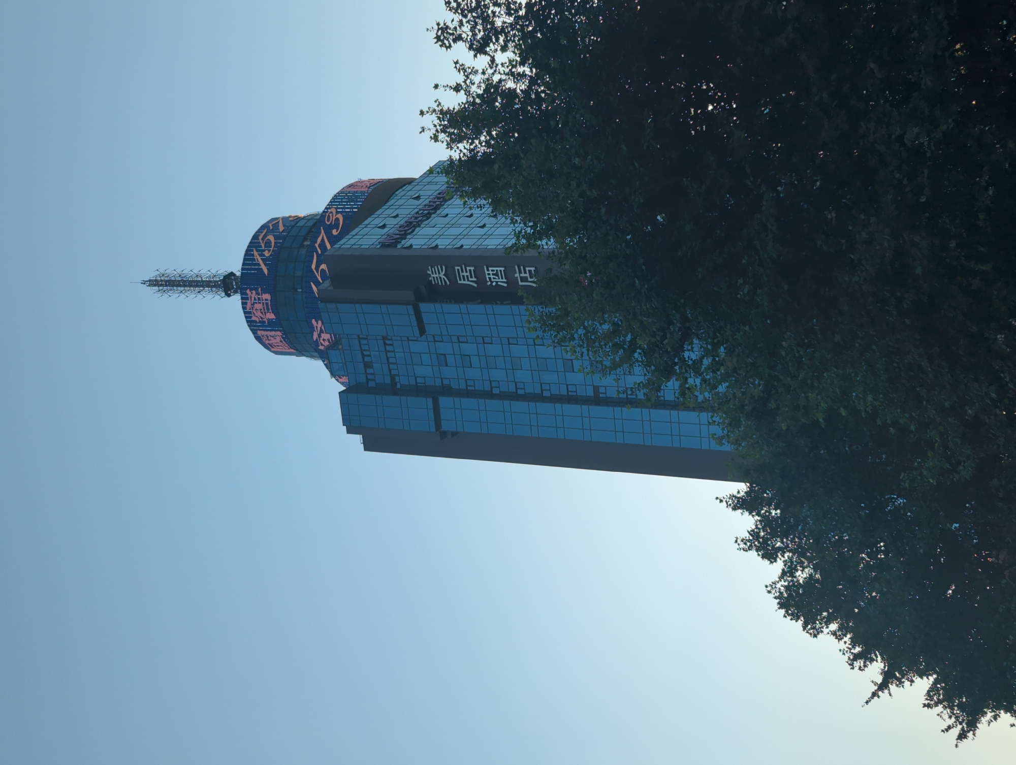 The image captures a close-up, low-angle view of the upper section of a tall, modern skyscraper in Jinjiang District, China. The building features a predominantly blue glass facade and is topped with a circular structure adorned with red illuminated text and numbers, including "1573". A telecommunications antenna is visible at the very peak of the building. To the right and bottom of the skyscraper, the dense foliage of green trees partially obscures the lower portions of the building. The sky is a clear, light blue. A vertical sign on the building's facade displays white Chinese characters that translate to "Meiju Hotel".