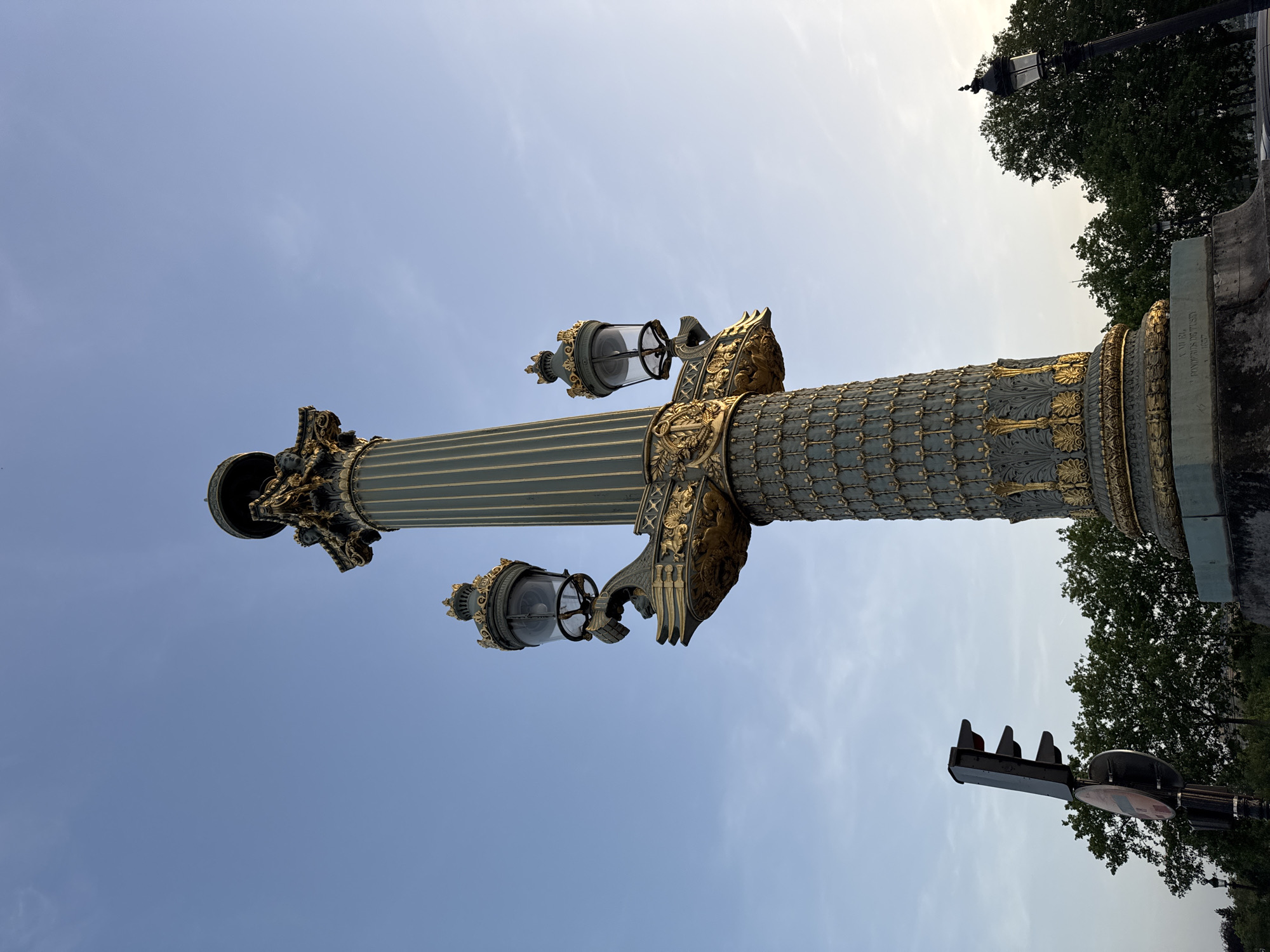 This image depicts a bronze and gold lamp post, located at the Place de la Concorde in Paris, France. The ornate lamp post features intricate detailing, including sculpted figures and decorative accents. The sky is visible in the background. Trees and traffic signals are to the right of the lamppost. The image appears to have been taken from a low angle, looking upwards.