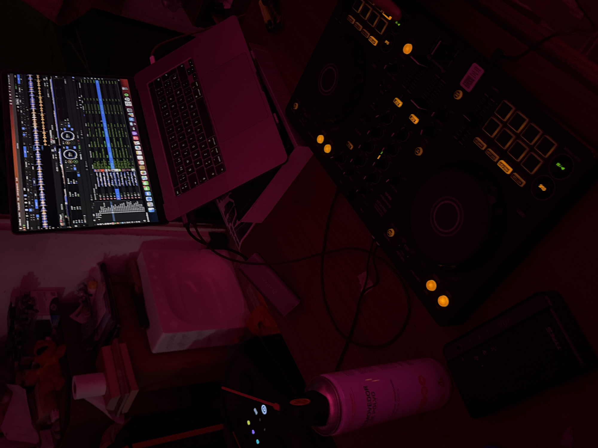The image captures a dimly lit, overhead view of a DJ setup on a desk, bathed in a strong pink light. No people are visible.
In the center-left, an open laptop (likely a MacBook Pro) displays DJ software, showing audio waveforms, track lists, and various application icons along the bottom dock, including Music, App Store, Settings, Safari, and Mail. To the right of the laptop is a DJ controller, possibly a Pioneer model, distinguished by its black surface and numerous illuminated yellow/orange buttons and knobs, suggesting it is powered on and active.
The desk surface around the equipment is cluttered with various items. Below the laptop, there's a stack of books, a roll of white paper (possibly toilet paper), and a small orange Elmo toy. To the right, next to the controller, lies a pink spray can labeled "REMOVEDOR DE POLVO" (Dust Remover) and "COLOUR". Further right, a black rectangular device with the text "THRIVES" is visible. Several cables snake across the desk, connecting the devices. A white, rectangular object (possibly foam packaging) is seen in the upper right quadrant of the image.
The scene suggests an indoor setting, likely at night or in a room with controlled lighting, given the dark surroundings and the glowing lights of the electronic equipment. The overall ambiance is moody and focused, indicative of a music production or DJing session taking place in Puebla, Mexico.