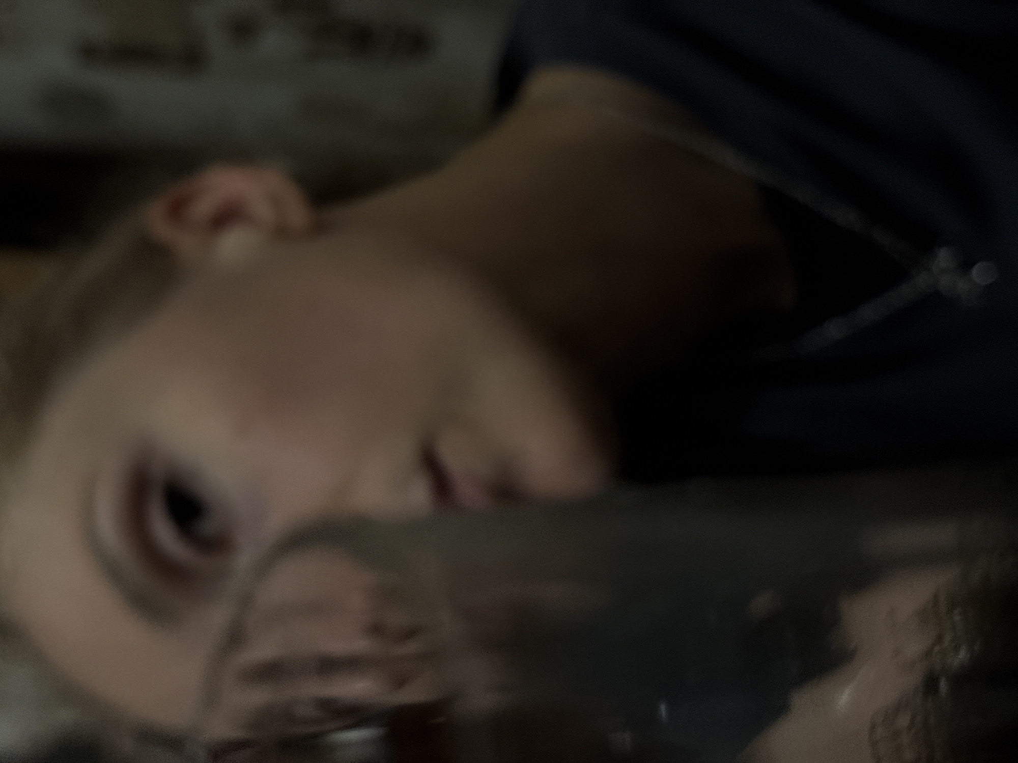 The image depicts a close-up view of a person lying on a surface, seemingly indoors. The person is fair-skinned, with blonde hair and appears to be wearing a necklace. The focus is soft, obscuring details. The background is dark and indistinct. There's no discernible context or information to suggest a location within Perm, Russia. The scene offers no indication of actions, interactions, or environmental details.