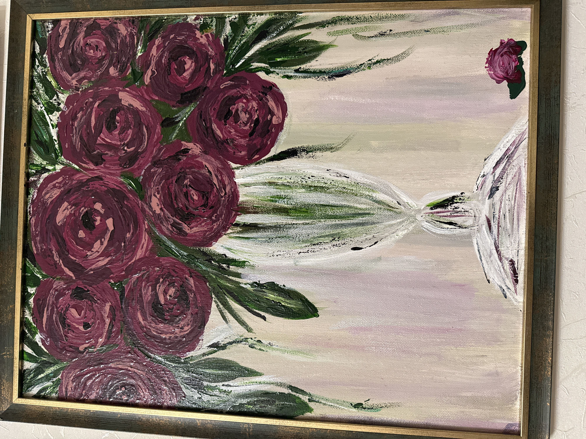 The image is a close-up, eye-level shot of a framed painting. The painting is an impressionistic depiction of a bouquet of dark red roses in a vase. The roses are rendered with thick, textured brushstrokes, and green leaves are visible behind and around them. The vase is white with vertical green and white lines, and it rests on a white, textured surface. The background of the painting is a soft, gradient of light pink, beige, and pale green. In the lower right corner of the painting, there is a single, small dark red rose and a green leaf. The frame of the painting has a dark green exterior and a gold inner edge. The painting is hanging on a textured, off-white wall. There is no indication of the city of Khabarovsk, Russia, or any specific location within it.