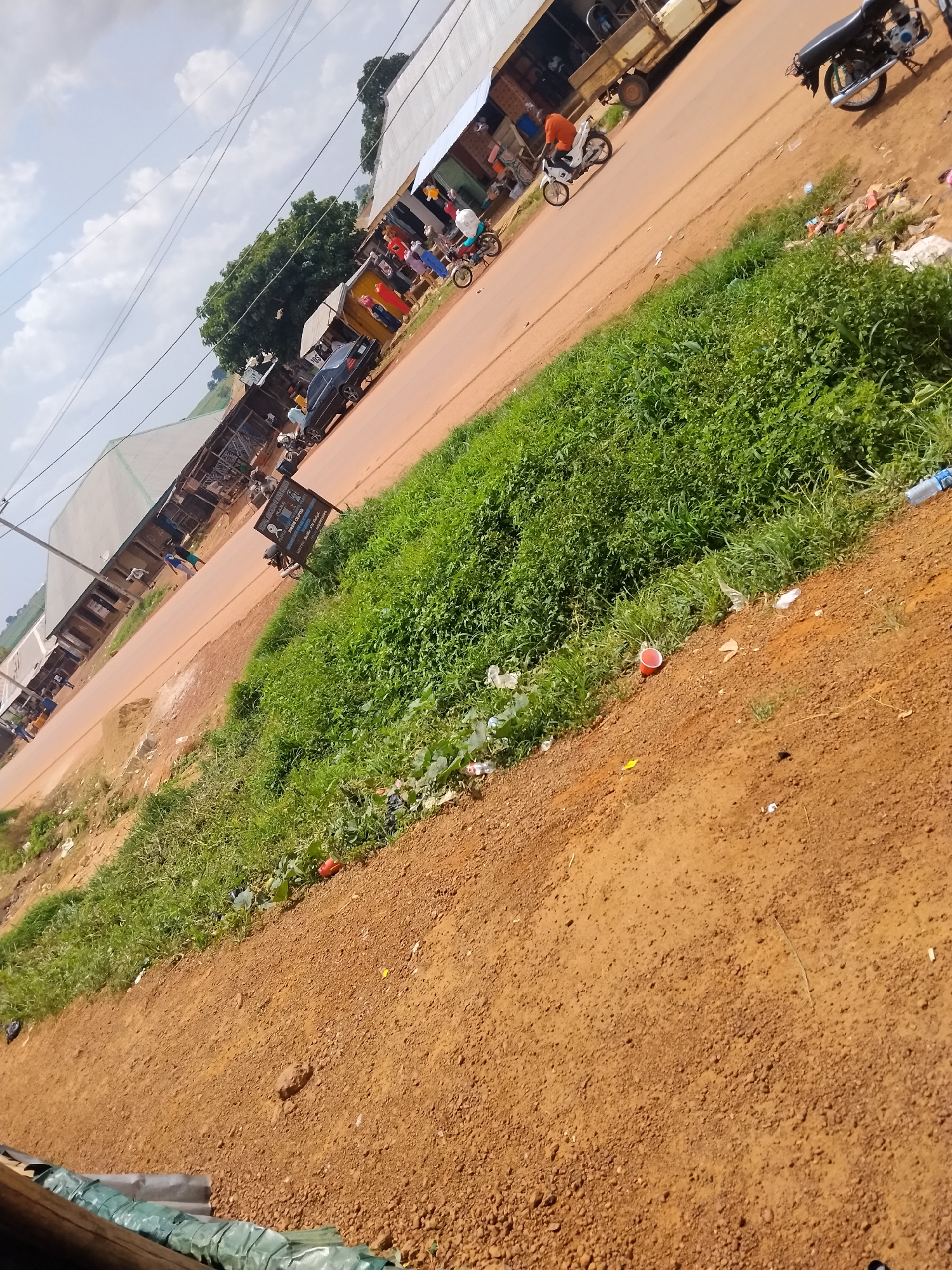 The image depicts a street scene in Durumi, Nigeria. The road is paved with reddish-brown soil. On the right side of the road, there is a patch of overgrown green grass, interspersed with litter including plastic bottles, red cups, and other debris. In the distance, along the left side of the road, are various market stalls or shops with corrugated metal roofs. People are visible walking along the roadside and some are gathered around the market stalls. Several motorcycles and a truck are parked or moving on the road. The sky above is partly cloudy.