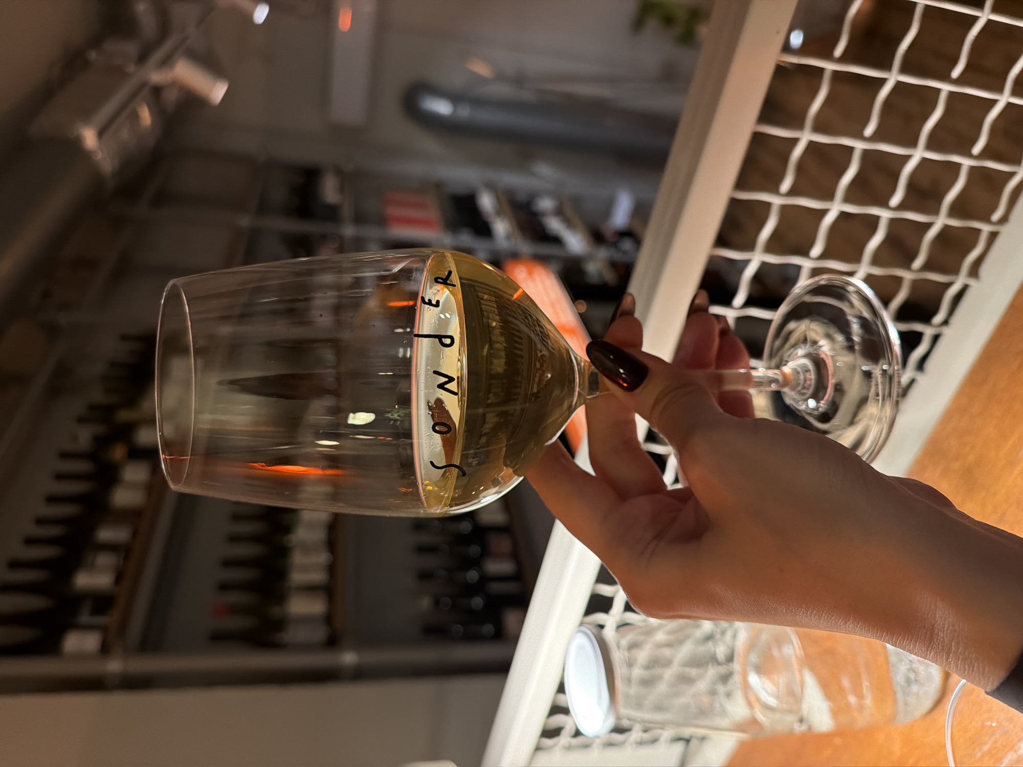 An indoor scene depicts a person's right hand holding a stemmed wine glass, approximately half-full with a light yellow liquid, likely white wine. The word "sonder" is visible on the glass. The individual's fingernails are painted with a dark, possibly plum or deep red, polish. The hand and glass rest above a light-colored wooden surface. To the left, a clear glass bottle with a silver cap, partially filled with water, is present.
In the background, a white metal grid railing spans horizontally across the lower mid-section. Behind the railing, the environment suggests a wine bar or restaurant, specifically identified as Sonder Wine Bar, located at Svetogorska 2, Belgrade, Serbia. To the left, a wall features multiple shelves stocked with numerous dark-colored wine bottles. To the right, a plain wall is visible, equipped with an air conditioning unit and overhead track lighting fixtures containing multiple spotlights. A tall, blurred, silvery, cylindrical object stands on the right side of the background. The overall lighting is dim and warm.