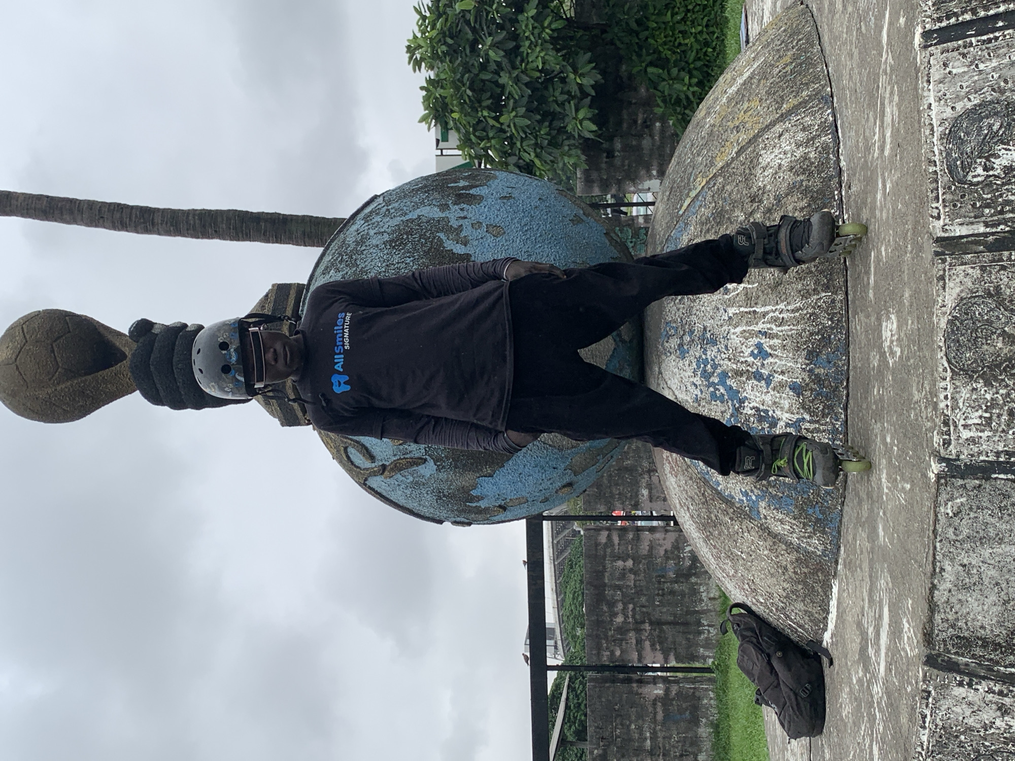A young Black male stands on a concrete platform wearing rollerblades, black pants, a black long-sleeved shirt with a blue "All Smiles Signature" logo on the left chest, and a black helmet. He is positioned in front of a large, weathered globe sculpture with peeling blue and tan paint. To his left, a black backpack rests on the platform. Behind him, a palm tree and lush green foliage are visible. The scene appears to be outdoors in an urban setting, likely in Iganmu, Nigeria, given the context. The sky is overcast.
