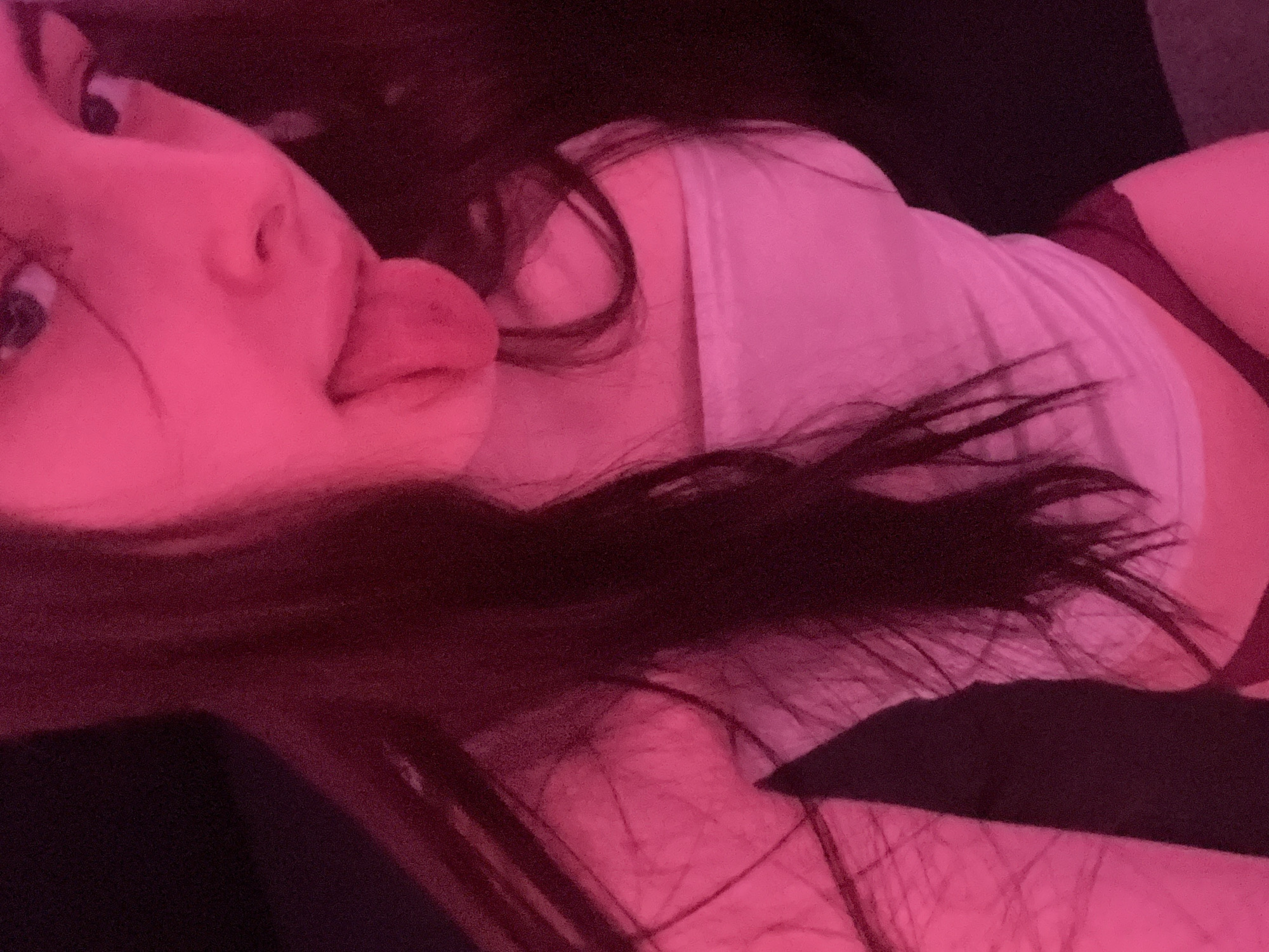 A person with long, dark hair is lying down, with their head tilted and tongue sticking out. They are wearing a light-colored t-shirt and dark-colored underwear or shorts. The image has a strong pink hue. The location cannot be determined from the image.