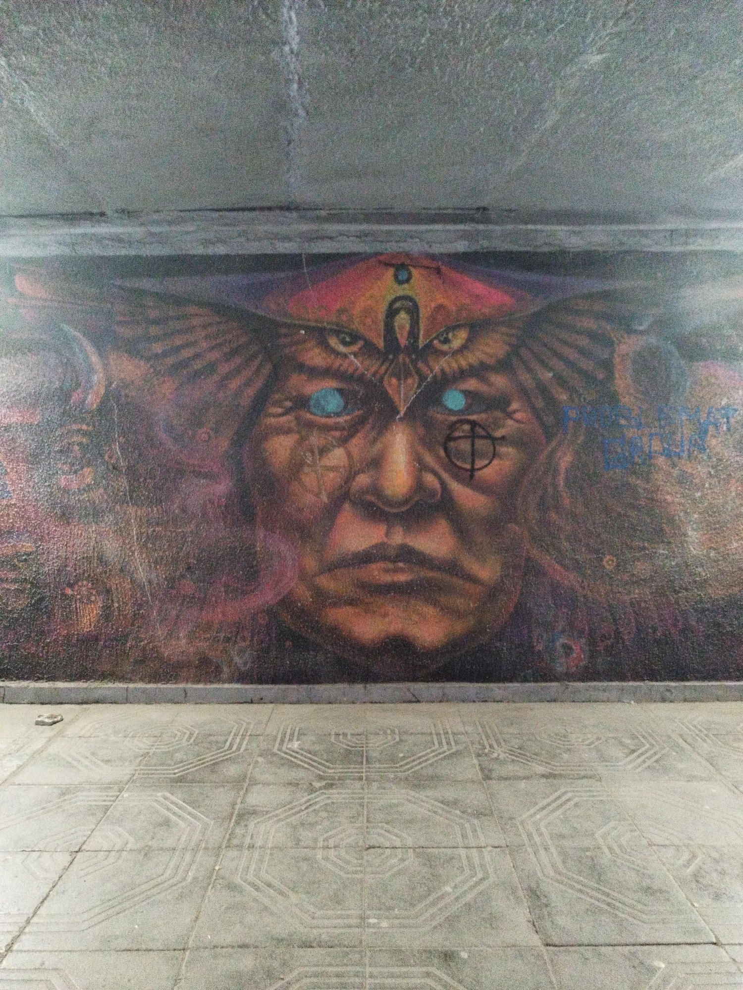 The image depicts a mural on a wall, likely inside a pedestrian underpass, in Sofia, Bulgaria. The mural features a large, stylized portrait of a face. The face is aged with blue eyes and a third eye above the brow, with feathered wings coming from the sides of the face. The background is composed of abstract shapes and colors. The floor in front of the mural is made of square tiles with a geometric pattern. The ceiling above the mural is concrete. There are no people visible in the image. Graffiti is visible on the upper right of the mural. A small rock is on the floor.