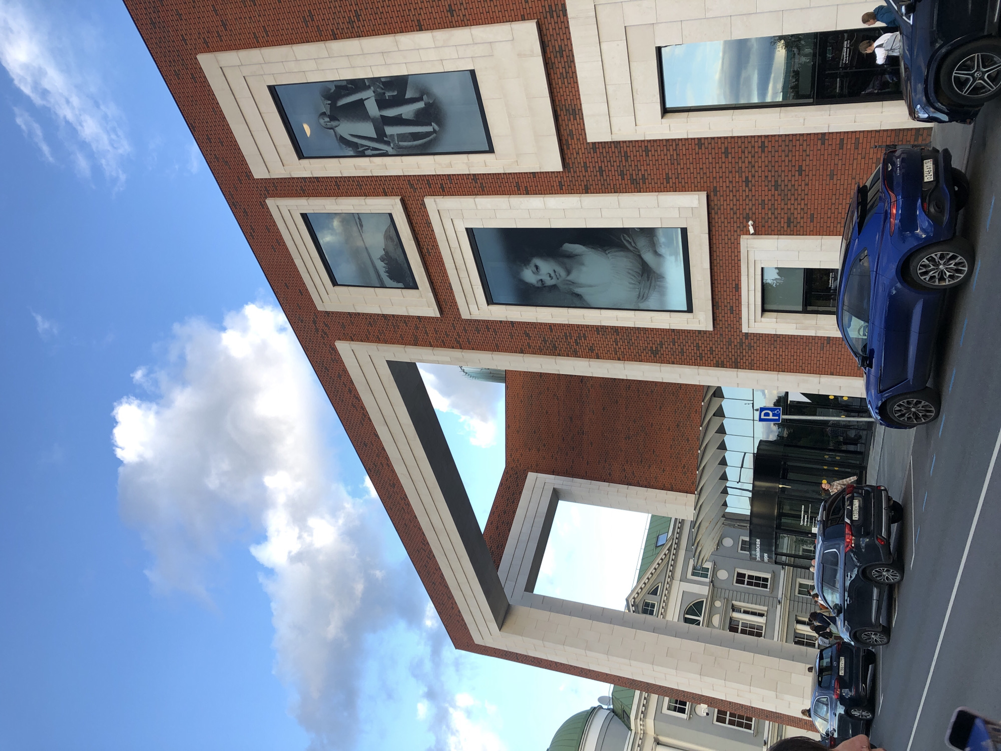 The image captures the exterior of the Garage Museum of Contemporary Art in Moscow, Russia. The museum's facade is a striking combination of red brick and large, minimalist white frames surrounding various artworks displayed behind glass. These artworks include a landscape, a portrait of a woman, and a figure in a wheelchair. In front of the museum, a blue Aston Martin DBX is parked on the street, alongside other vehicles. The street in front of the museum is lined with trees and has a sidewalk. The sky above is a mix of blue and white clouds. The overall scene suggests a contemporary urban environment with a focus on art and architecture.
