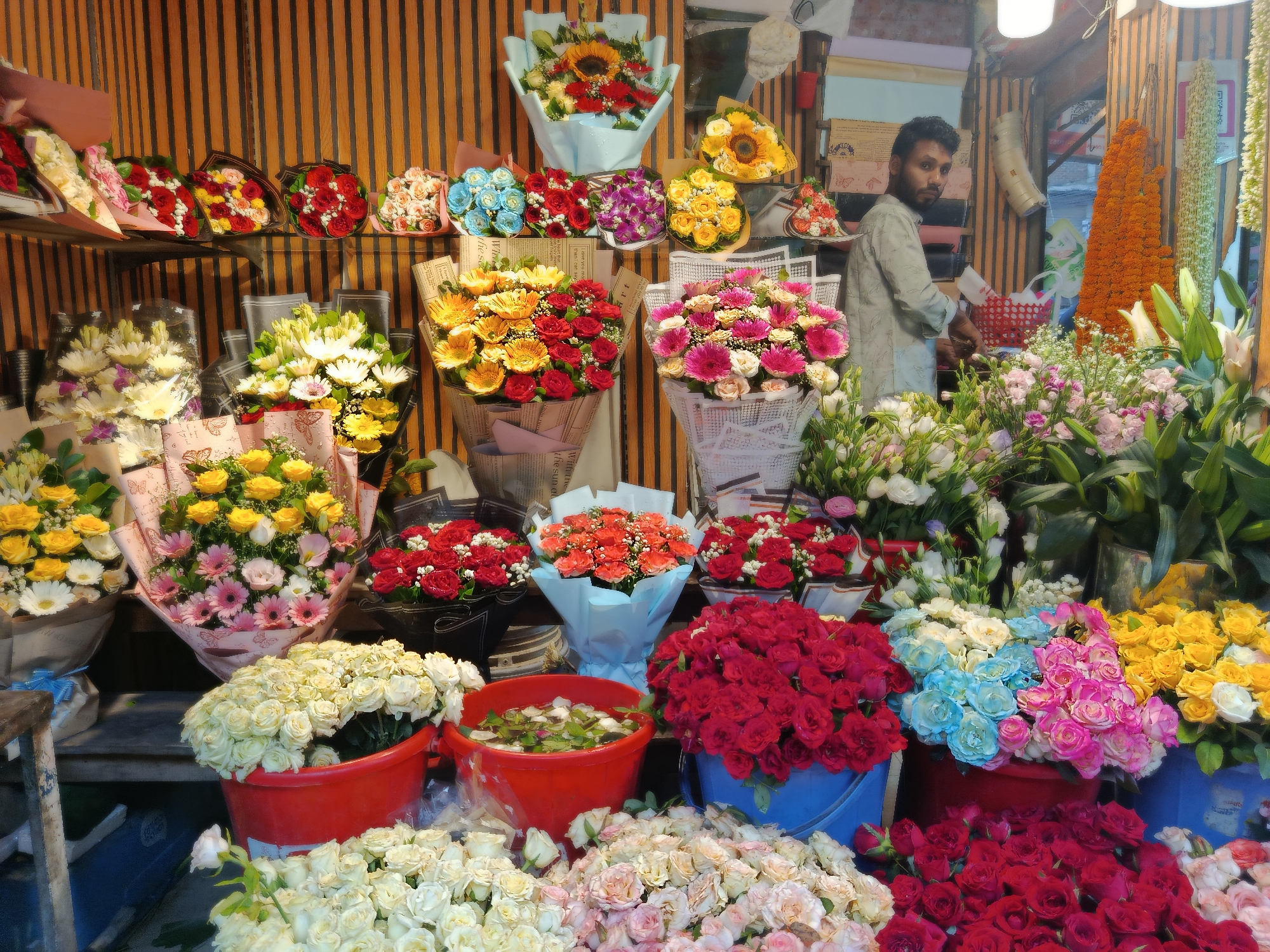 The image showcases a vibrant flower shop, likely located in Dhaka, Bangladesh. A man is visible inside the shop, amidst a colorful and diverse array of floral arrangements. Bouquets of sunflowers, roses, daisies, and other blossoms are arranged on shelves and displayed in buckets, presenting a profusion of color and texture. Garlands, strung up in the background, suggest the shop might be catering to events or traditional celebrations. The setting indicates a bustling market environment.