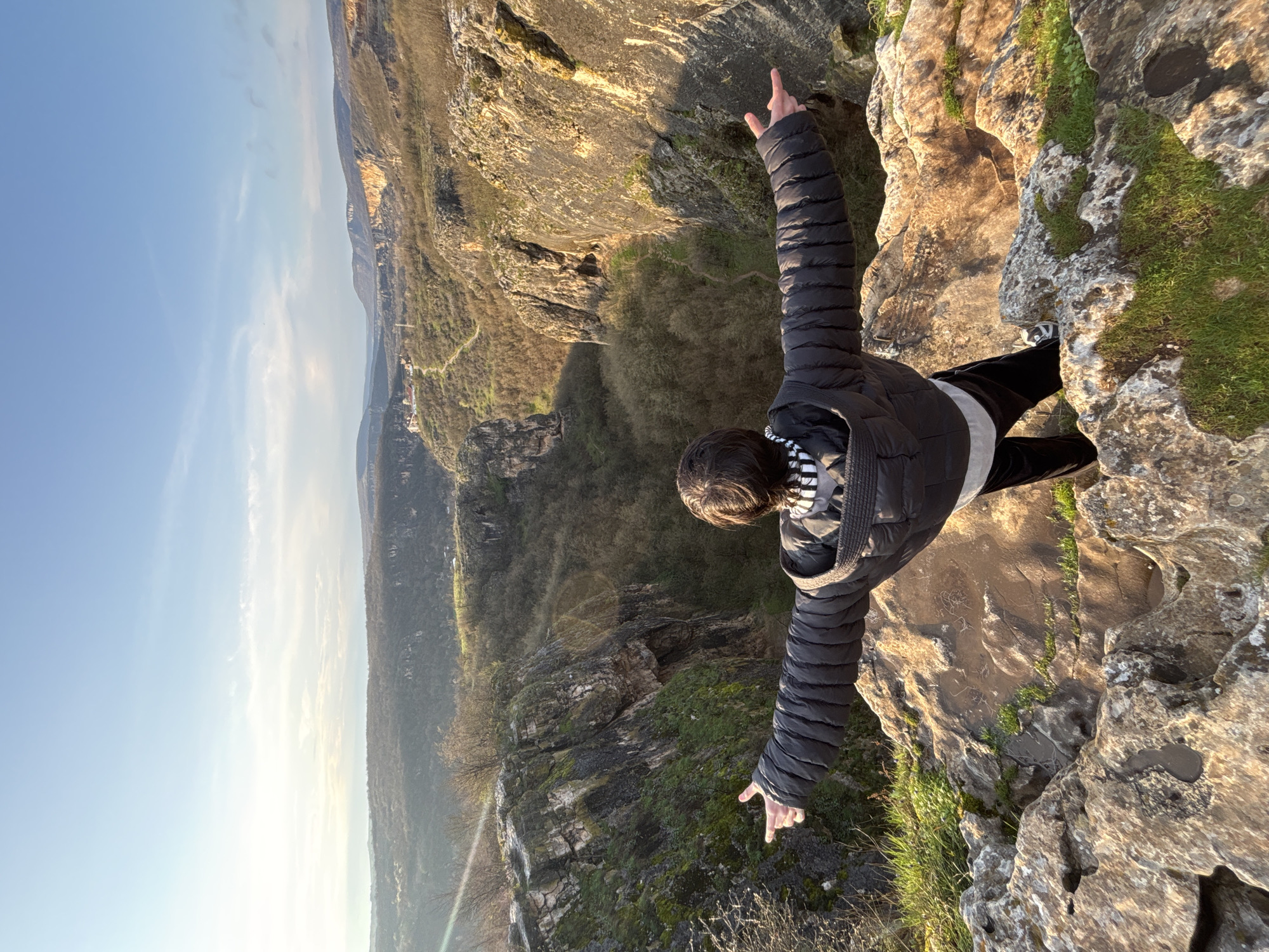 A person stands on a rocky precipice overlooking a valley, with their arms outstretched to either side. The person wears a dark, quilted jacket over a white top with a striped collar, and dark pants. The rocky surface is covered with patches of green grass and moss. Below, a dense forest fills the valley floor, with cliffs and rock formations visible through the trees. In the distance, a blue sky stretches over a landscape of rolling hills. The location is likely in the vicinity of Karlukovo, Bulgaria, given the karst topography typical of the area.