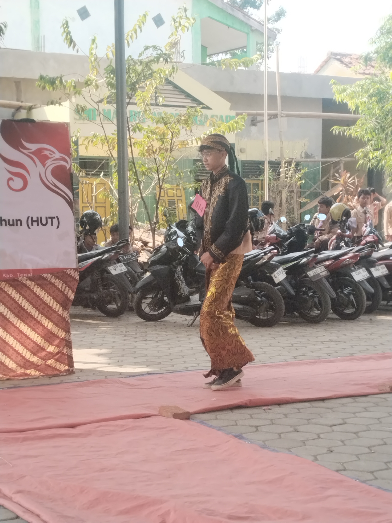 In Pakulaut, Indonesia, a young man wearing traditional Javanese attire, including a black embroidered jacket and a gold patterned sarong, stands on a pink tarp in front of a line of parked motorcycles. He has a dark head covering and glasses. Behind him, several other young people, some in school uniforms, are gathered, and a banner with a red eagle design and text is visible on the left. The scene appears to be an outdoor event, possibly a cultural festival or competition, given the presence of the banner and the participant's attire. The environment includes buildings with light-colored walls and green roofs, trees with sparse foliage, and a paved ground surface.