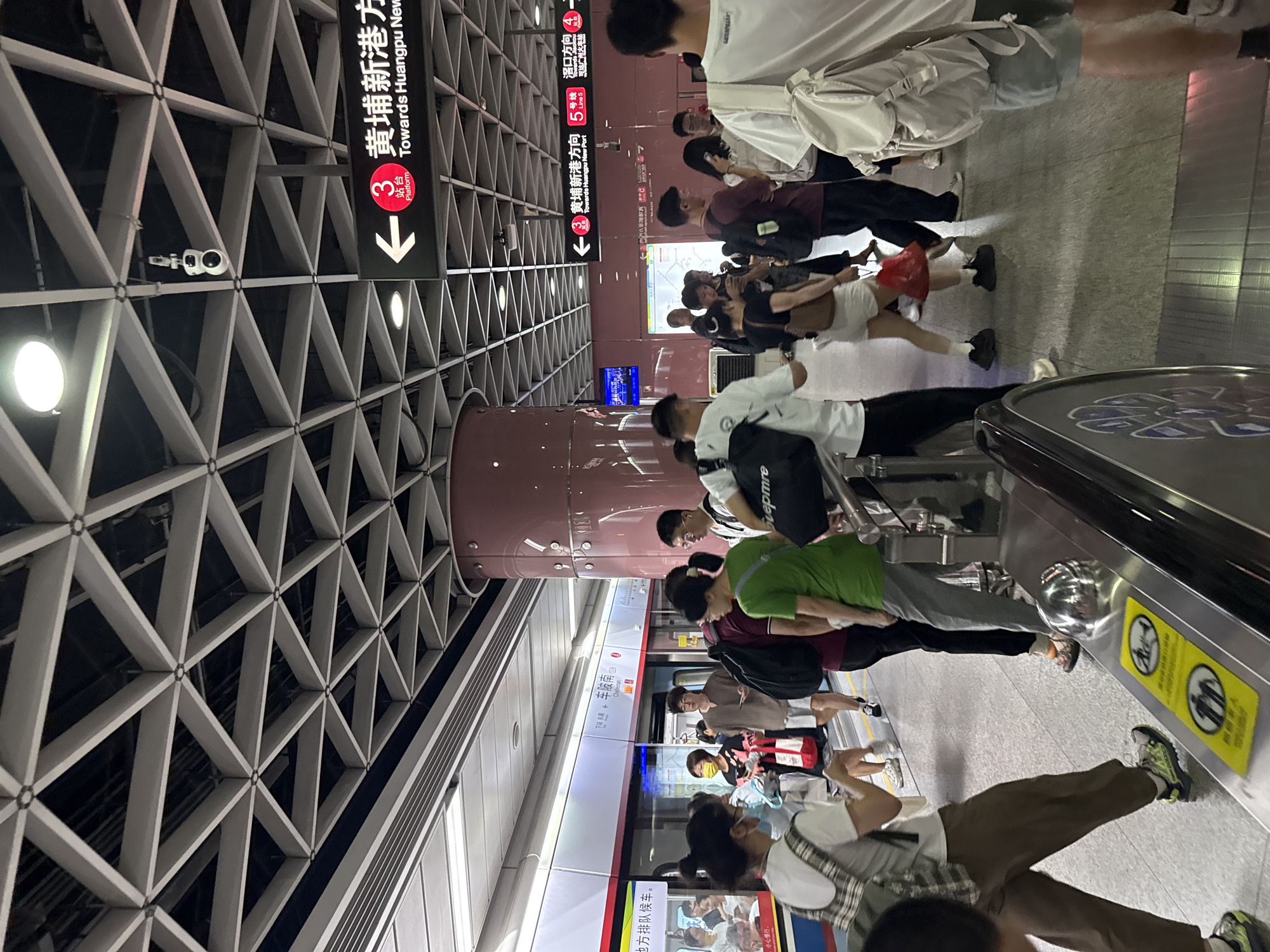 The image depicts a busy subway platform in Tianhe District, China. Numerous people, predominantly young adults, are gathered along the platform, waiting for a train. The scene captures a typical commuter environment, with individuals standing, some looking at their phones, and others conversing.
The station's interior features a distinctive white geometric lattice ceiling structure with integrated lighting fixtures. Large, reddish-brown cylindrical pillars are spaced along the platform. A subway train, primarily white with a red stripe, is present at the platform, its doors closed. Part of an escalator or travelator with a patterned surface is visible in the foreground on the right.
Prominent signage in both Chinese and English indicates directions and line information. A large overhead sign points "Towards Huangpu New Port" for Platform 3, showing "3 黄埔新港方向" and "站台" (Platform). Another sign confirms "←③ 黄埔新港方向" (Towards Huangpu New Port) and mentions "5号线" (Line 5), stating it "可达广州火车站" (Can reach Guangzhou Railway Station). A small red square with "4" is also visible. The train's digital display shows "车陂南" (Chebenan). Floor stickers with yellow circular backgrounds contain symbols and Chinese characters like "小心慢行" (Walk carefully) and a "no running" icon. Text visible on clothing includes "Svepmre" and "CATERPILLAR". A white security camera is mounted on the ceiling structure. The overall lighting suggests it is daytime or early evening.