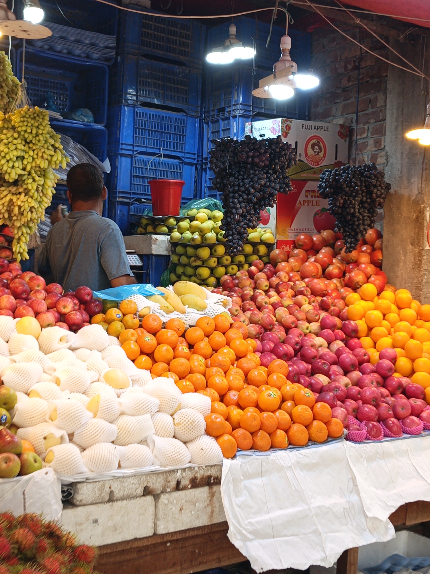 The image depicts a vibrant fruit stall in Dhaka, Bangladesh. A vendor, facing away from the camera, stands behind a table laden with diverse fruits, including green grapes, black grapes, apples, oranges, pears, guavas and tangerines. The guavas are wrapped in white protective mesh. A red bucket sits atop a stack of green apples. The fruit is arranged in mounds. Behind the vendor, blue plastic crates are stacked. Packaging for Fuji apples is visible. The stall is lit by overhead fluorescent and incandescent bulbs. A brick wall is visible.
