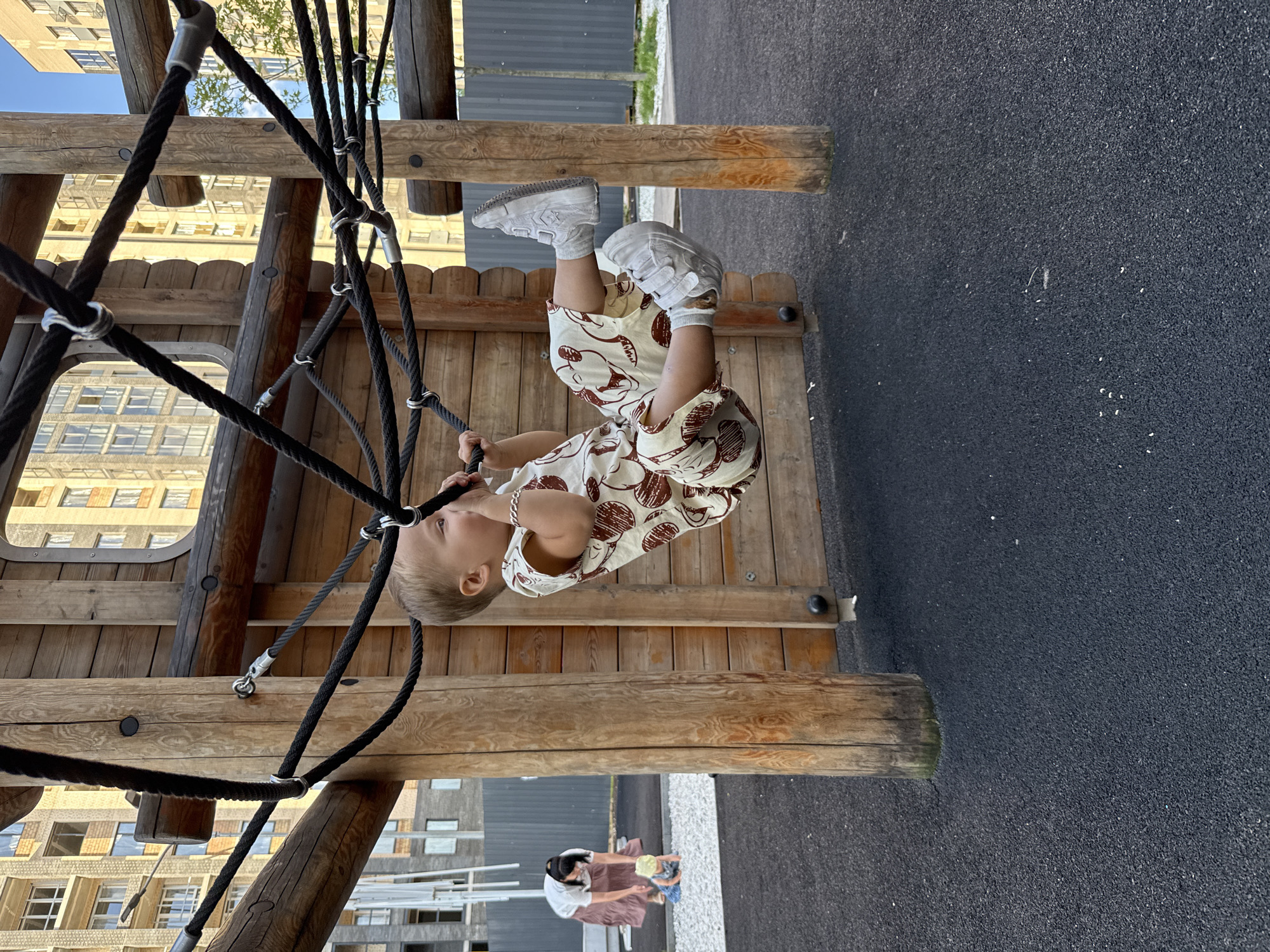 In Rusanovo, Russia, a child is captured mid-air, holding onto black ropes of a wooden playground structure. The child wears a light-colored jumpsuit with a brown pattern of what appears to be cartoon bears and a silver bracelet. White sneakers are on their feet. In the background, a woman with dark hair, wearing a white shirt and a long, patterned skirt, kneels near another child who is on the ground in a fenced-off area with white gravel. The playground is surrounded by dark, speckled rubber ground cover. Behind the playground, modern multi-story buildings are visible, indicating an urban setting. The sky above is bright, suggesting daylight.