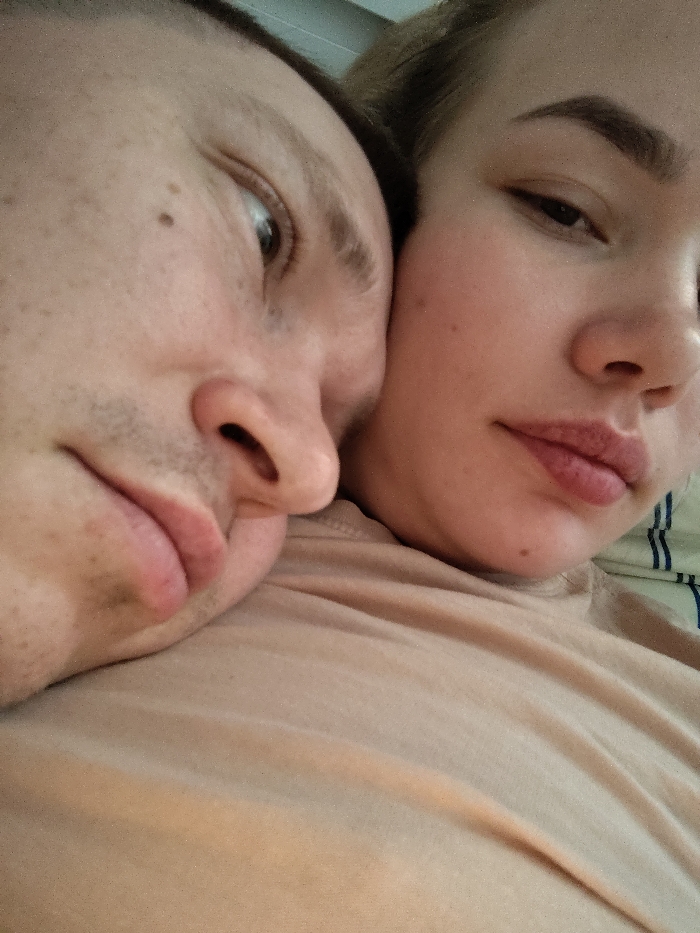 An overhead close-up image captures two individuals in an indoor setting in Марковский, Russia. The individual on the left is a male with fair skin, minimal facial hair (stubble on chin and upper lip), and a visible mole on his left cheek. His right eye is partially open, gazing towards the upper right. The individual on the right is a female with fair skin, light-colored hair, groomed eyebrows, and light lip color. Her left eye is open and directed towards the viewer. The faces of both individuals are in direct contact, with the male's left temple and cheek pressed against the female's right cheek and shoulder area. Both individuals are wearing solid light brown or tan-colored garments, appearing to be t-shirts. A striped fabric, possibly a pillow or sheet, is visible behind the female's head on the far right.