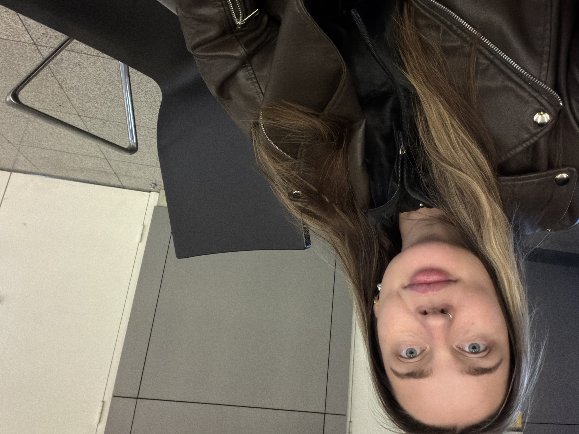 A woman with blonde-streaked brown hair, blue eyes, and fair skin is seen from an overhead angle. She has a silver nose ring in her left nostril and a gold earring in her left ear. She is wearing a brown leather jacket over a black shirt. She is seated in a black chair. The background consists of gray and white tiled walls and flooring. The location is likely a building interior in an urban environment, potentially in the city of Khimki, Russia.