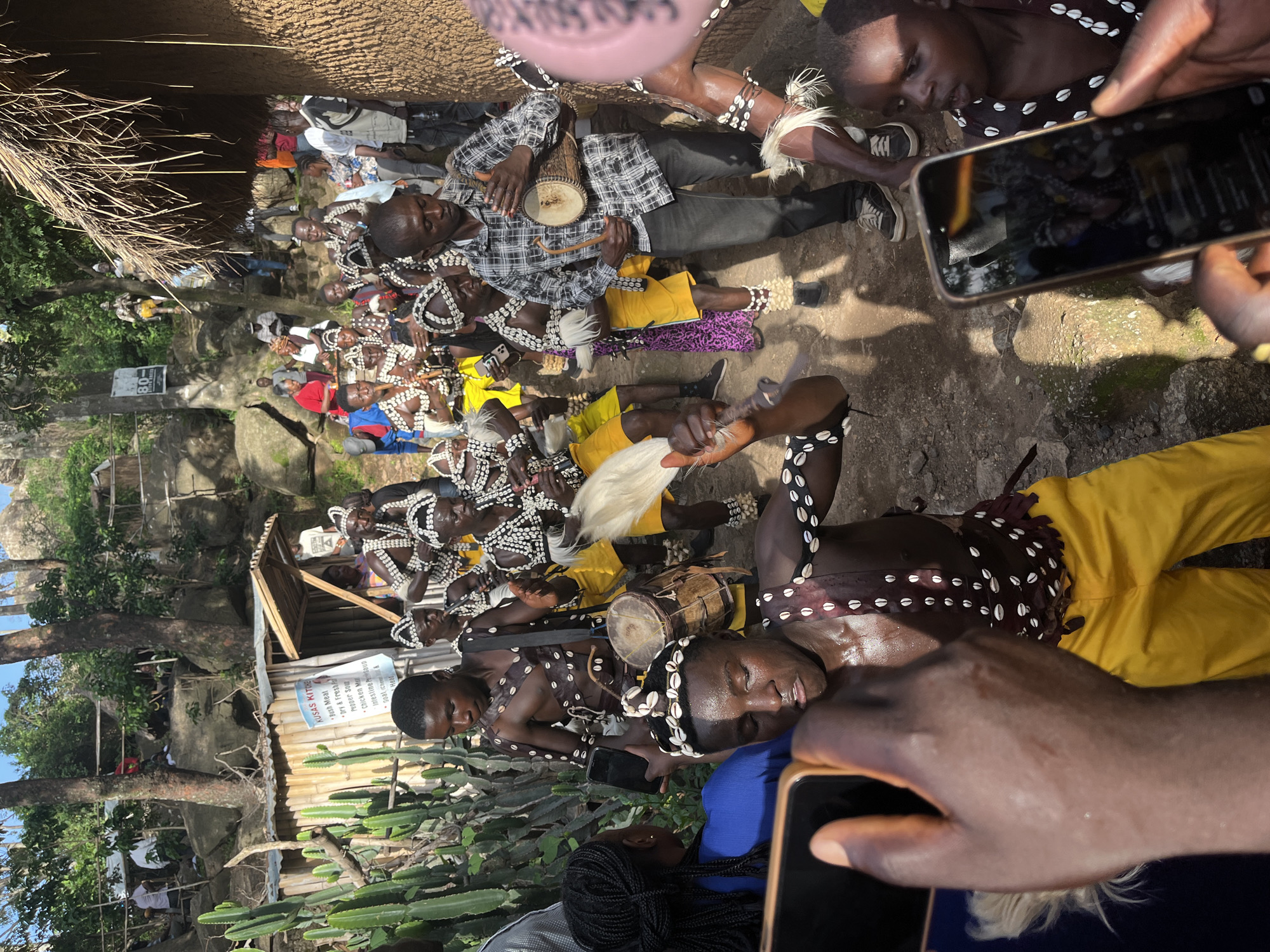This media file captures a gathering of people, likely during a cultural event or ceremony, in what appears to be an outdoor, somewhat rustic setting in Jos, Nigeria. The foreground is dominated by the hands and upper bodies of several individuals, some of whom are dressed in traditional attire. This clothing features intricate beadwork and shell adornments around the neck, wrists, and head.
Several people in the image are playing musical instruments, including drums and what seems to be a stringed instrument. One person is seen holding a small drum and striking it with a stick. Another individual appears to be blowing into a pipe or flute. The atmosphere suggests a lively occasion, possibly a celebration or ritual.
In the background, more people are visible, some seated and observing, while others seem to be participating in the event. There are also elements of the natural environment, with trees and rocky formations suggesting a natural or semi-natural landscape. A wooden structure with a corrugated metal roof is also present, possibly a stall or a small building.
Visible text on a sign in the background reads "KUSAS KITCHE" and lists various food items like "Bush Meat," "Pepper Soup," and "Chicken." This indicates the presence of a food vendor or establishment at the event. The time of day appears to be daytime, with natural light illuminating the scene. The overall mood seems to be one of community engagement and cultural expression.
