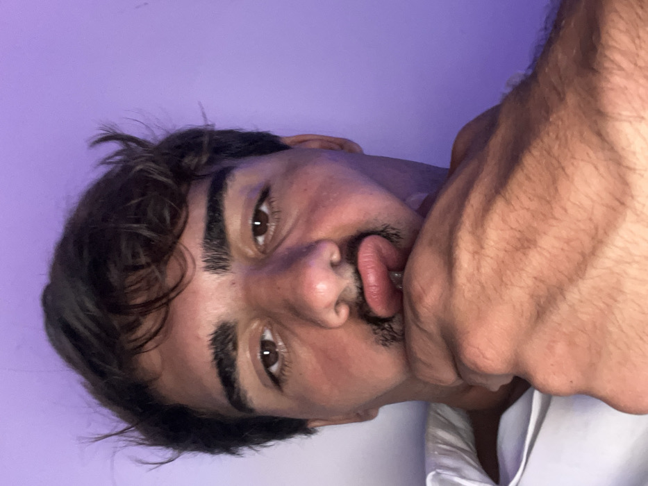 A young man with dark, curly hair and thick eyebrows poses close to the camera with a purple-toned background. His brown eyes stare directly at the viewer. His tongue protrudes from his mouth and he holds his hand closely over his mouth and nose, partially obscuring his face. His shirt, visible at the bottom edge of the frame, is white. The image is rotated sideways, with the man appearing tilted.