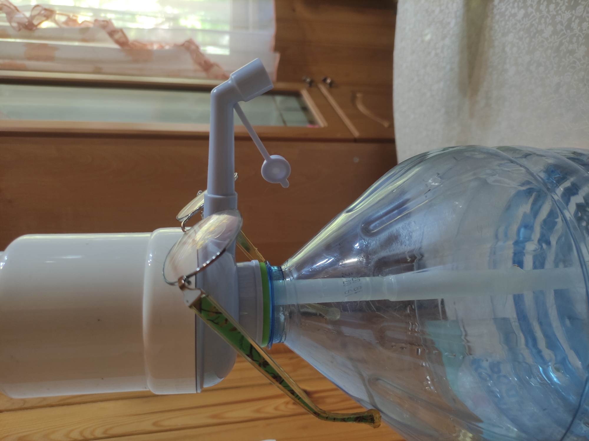The image shows a water bottle with a dispenser pump attached to its opening. A pair of eyeglasses with camouflage frames is perched on the dispenser. The setting appears to be indoors, on a wooden surface. The background features a wooden wall or cabinet, and a window with a light-colored curtain. The water bottle is partially filled with water. There is no indication of the location being Kratovo, Russia. There are no people depicted in the image.