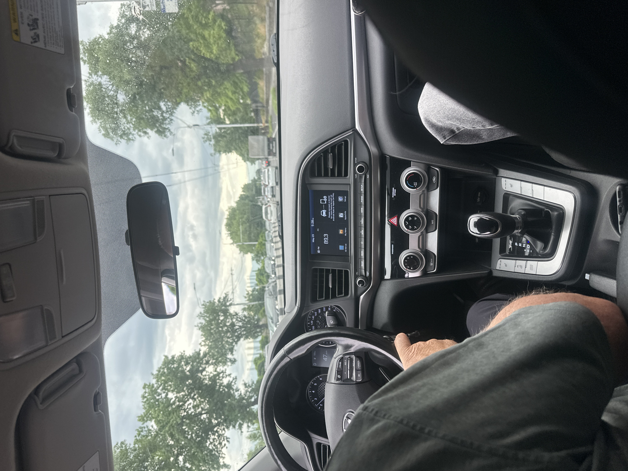 A view from inside a car, showing the dashboard, steering wheel, and part of the driver. The car is moving at 47 km/h. The radio is tuned to 89.3 FM, and the date on the screen is May 23. Outside the car, there are trees and parked cars. The location is not identifiable as being in Yerevan, Armenia from this image.