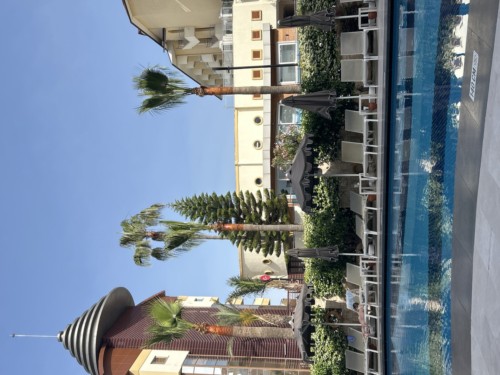 The image shows a sunny scene at a pool in what appears to be a resort. Palm trees are visible, as well as lounge chairs and umbrellas next to the pool. The pool itself is blue and reflects the surrounding buildings and foliage. The buildings in the background include a modern structure with a distinctive layered conical roof, as well as other hotel-style buildings. The scene suggests a tourist location, typical of Alanya, Turkey.