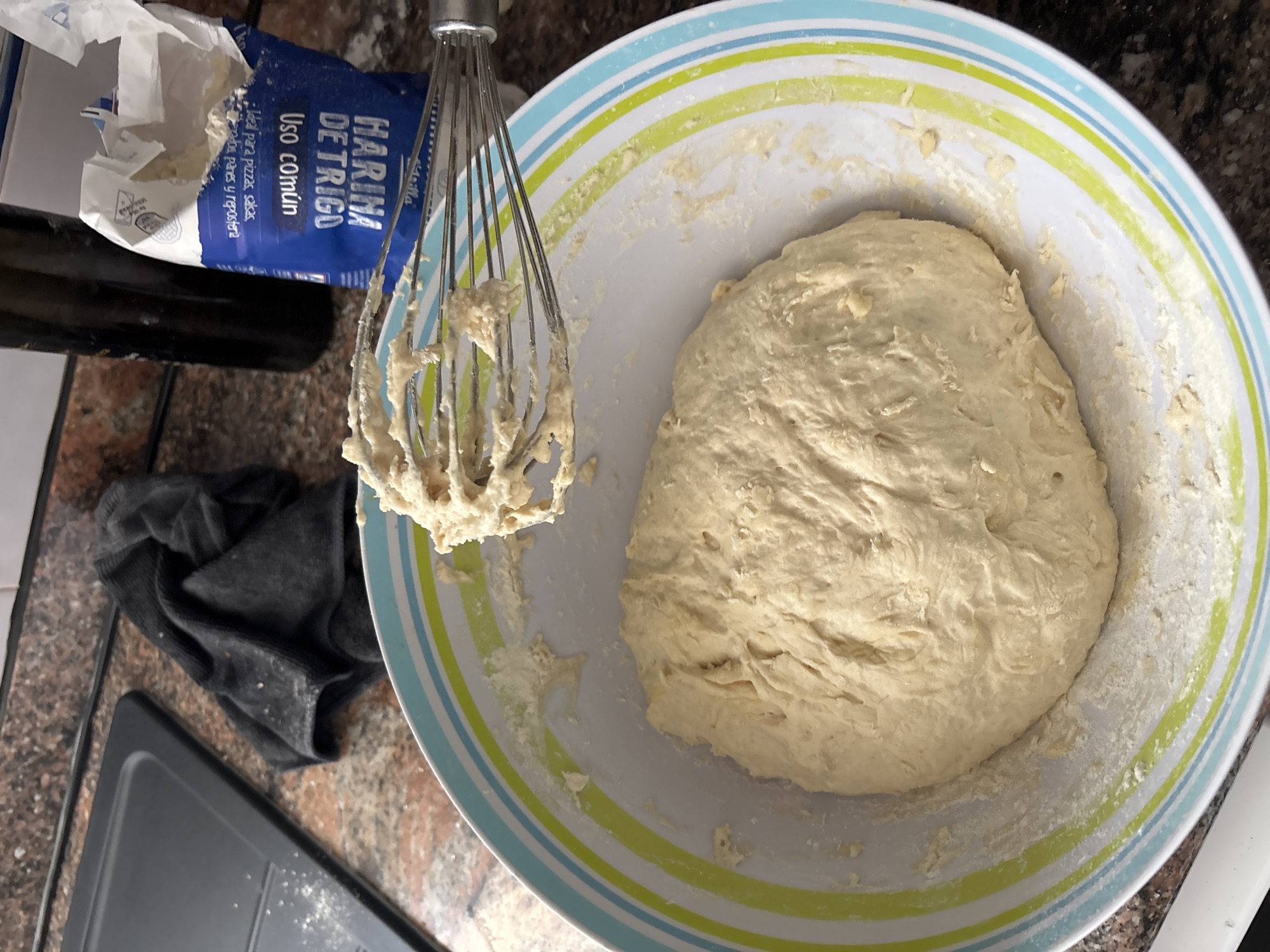 The image shows a close-up view of dough being prepared in a bowl. The bowl has blue, green, and white stripes around the rim. A metal whisk, coated with dough, is held above the bowl. A bag of "HARINA DE TRIGO USO COMUN" (all-purpose wheat flour) is visible in the background, along with a dark bottle. A black cloth is folded to the left of the bowl. The scene appears to be indoors, likely in a kitchen, with a countertop visible. There are no people or animals present. The lighting suggests it is daytime. The image does not provide any information about the location in the city of Orihuela, Spain.