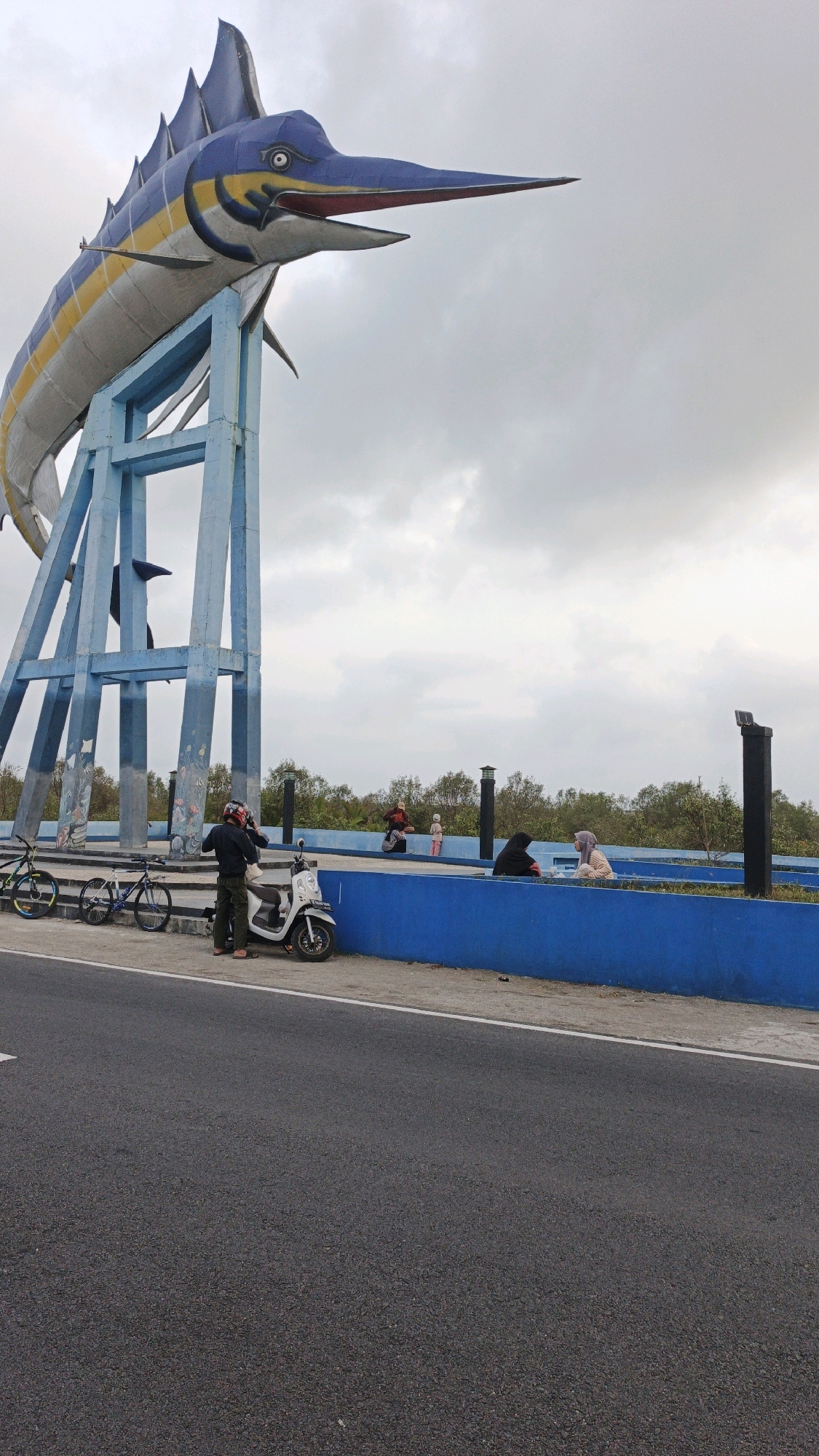 The image shows the Marlin Statue in Pangandaran, Indonesia. The statue is a large sculpture of a marlin fish mounted on a blue metal structure. In the foreground, a man wearing a helmet stands next to a white scooter. Two bicycles are parked nearby. In the background, several people are sitting on a blue wall. The sky is overcast with clouds. The scene appears to be a public area near the coast, given the presence of mangrove trees in the distance and the implied proximity to water.