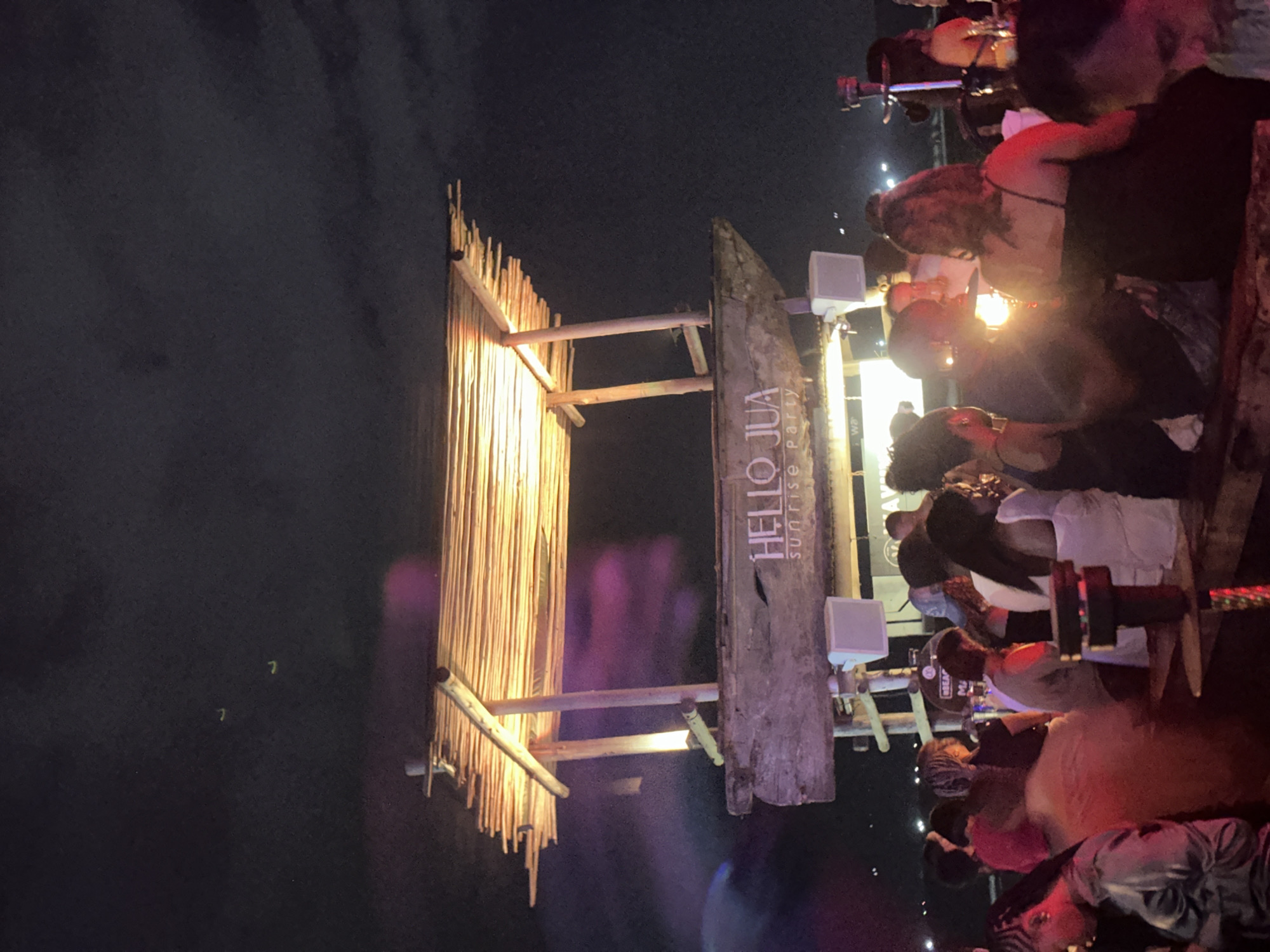 The image shows a nighttime scene, likely a party or event, with a crowd of people gathered around a structure. The structure appears to be constructed from wood and natural materials, possibly bamboo or reeds, and has the words "HELLO JUA sunrise Party" written on it. The location is in Unknown, Unknown. The crowd consists of a diverse group of people, some standing, some sitting, and some interacting with each other. There are lights illuminating the structure and the people around it, creating a festive atmosphere. The background is dark, suggesting the event is taking place outdoors at night.
