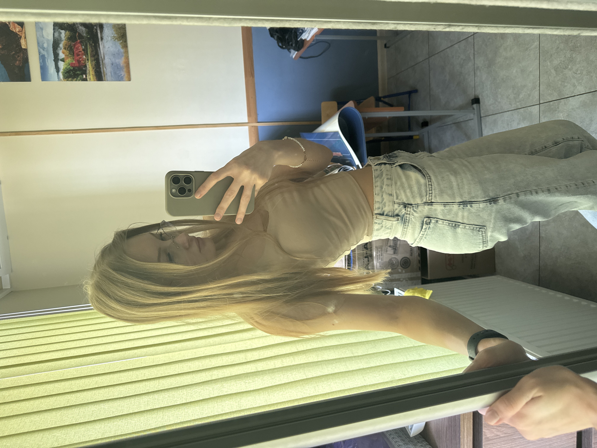 A fair-skinned young woman takes a mirror selfie. She wears light-wash denim jeans and a beige tank top. She has blonde hair, glasses, and a small nose ring. She is holding a phone with a light green case. The reflection shows a room with paintings on the wall, a desk, and a tiled floor. Blinds are in the foreground. The city is not visible and therefore location in Perm, Russia cannot be confirmed.