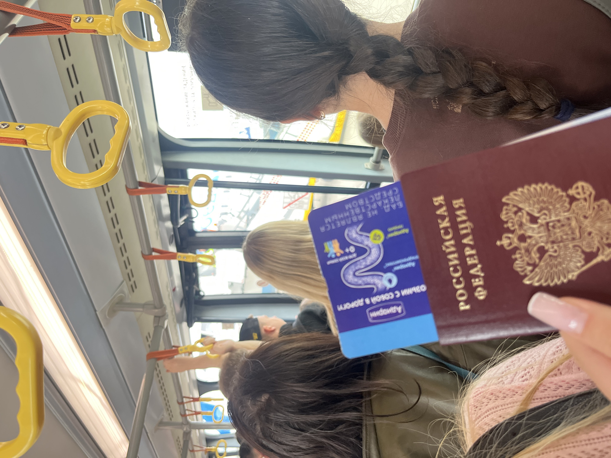In this medium shot, taken vertically, a person holds a Russian passport and a blue health supplement packet in a public transportation setting, likely a bus or train. The passport, with its distinctive Russian emblem, is in focus in the foreground, held by a hand with neatly manicured, light pink nails. To the left of the passport is a small, rectangular blue packet featuring a stylized purple snake graphic and text in Russian. The text on the packet includes "Адиорин", "ОЗЬМИ С СОБОЙ В ДОРОГУ!" (Take with you on the road!), and "БАД. НЕ ЯВЛЯЕТСЯ ЛЕКАРСТВЕННЫМ СРЕДСТВОМ" (Dietary supplement. Not a medicine).
The background reveals a view through a window showing a blurred urban landscape, suggesting the vehicle is in motion. Inside the vehicle, yellow handrails are visible, a common feature in public transport for passengers to hold onto. The lighting suggests it could be daytime. The subjects in the background are out of focus, but glimpses of people with dark and blonde hair can be seen, their faces not clearly discernible. The overall mood is candid, capturing a moment of travel preparation or journey. The presence of a passport and a health supplement implies travel, possibly international or a long-distance domestic trip. The setting appears to be Vnukovo, Russia, as indicated by the context.