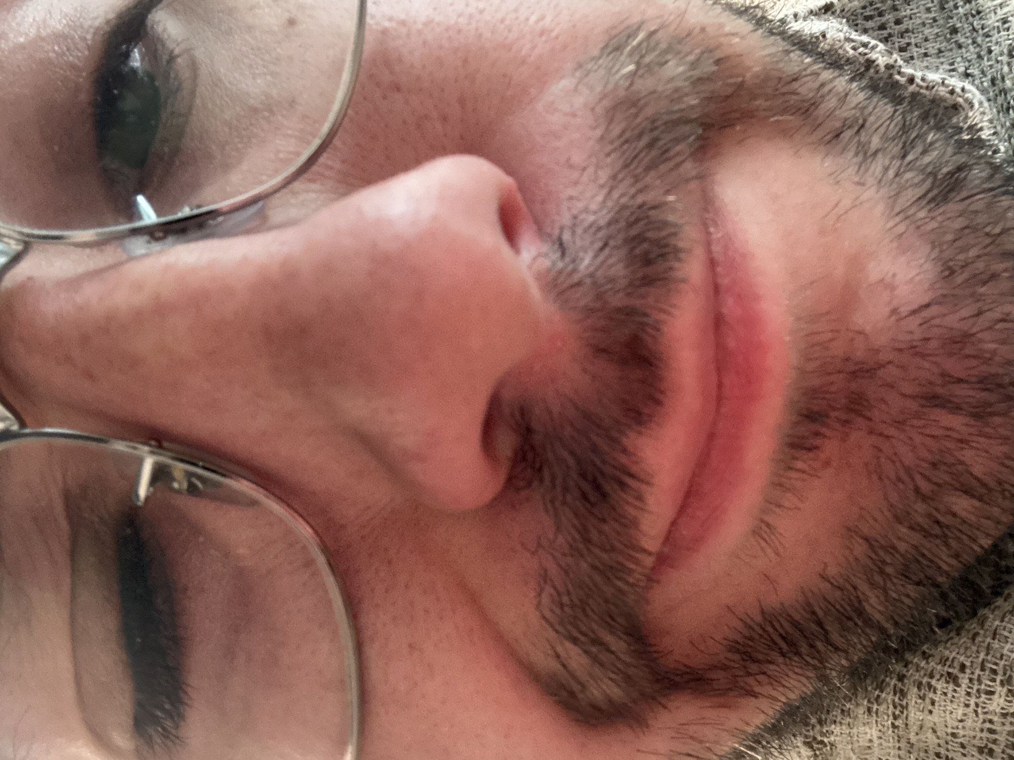A close-up, upside-down image shows a portion of a person's face. Visible features include an eye with glasses, a nose, a mouth with a mustache, and a portion of their head resting on what appears to be a textured fabric. The skin tone is light. The person's facial hair is dark. The location is not discernible from this image.