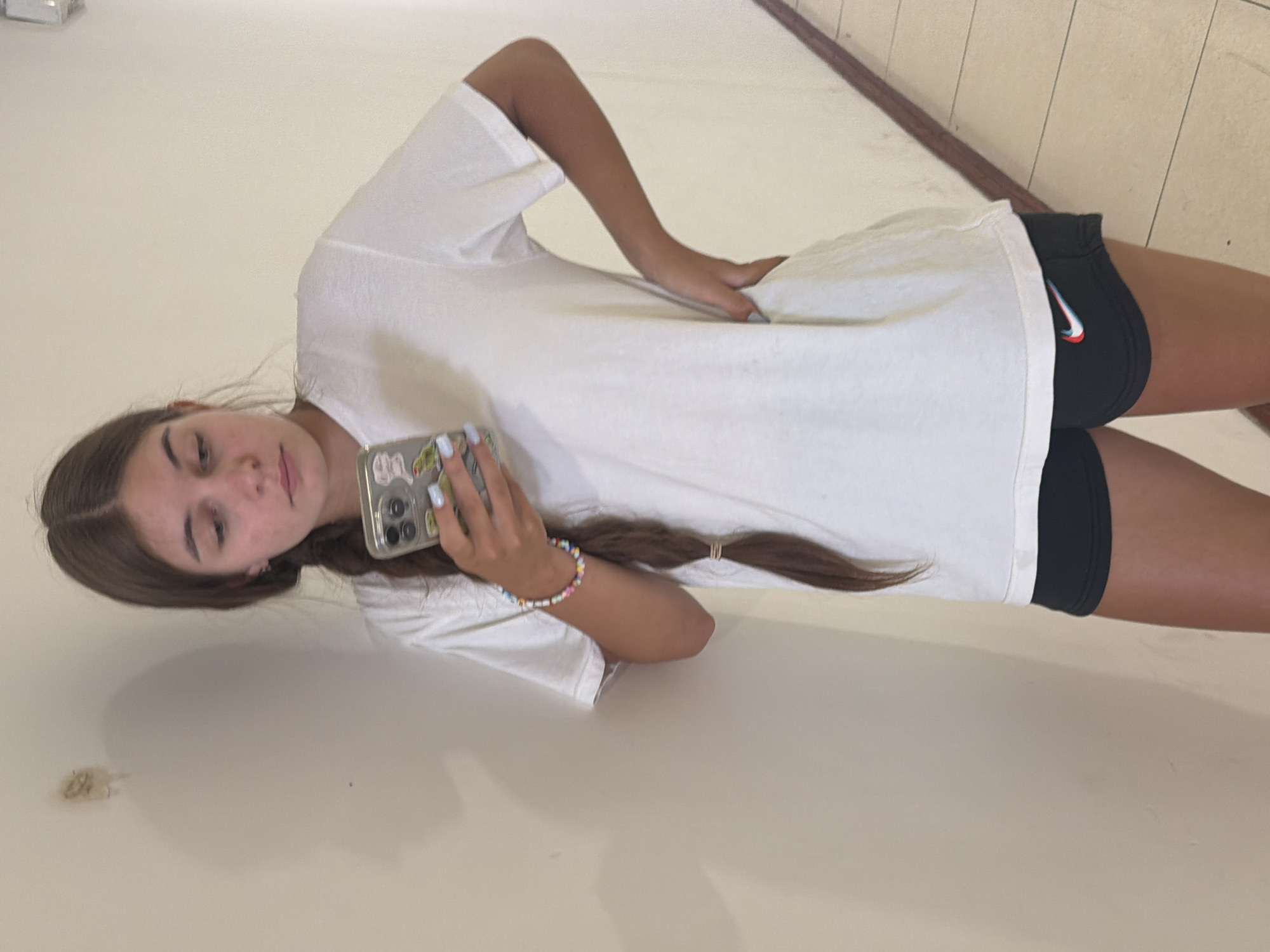 A young woman with brown hair in a braid and a white t-shirt and black shorts stands in a hallway in Nha Trang, Vietnam. She is holding a smartphone in her left hand and has her right hand on her hip. The hallway has a light-colored wall and a tiled floor with a wooden trim.