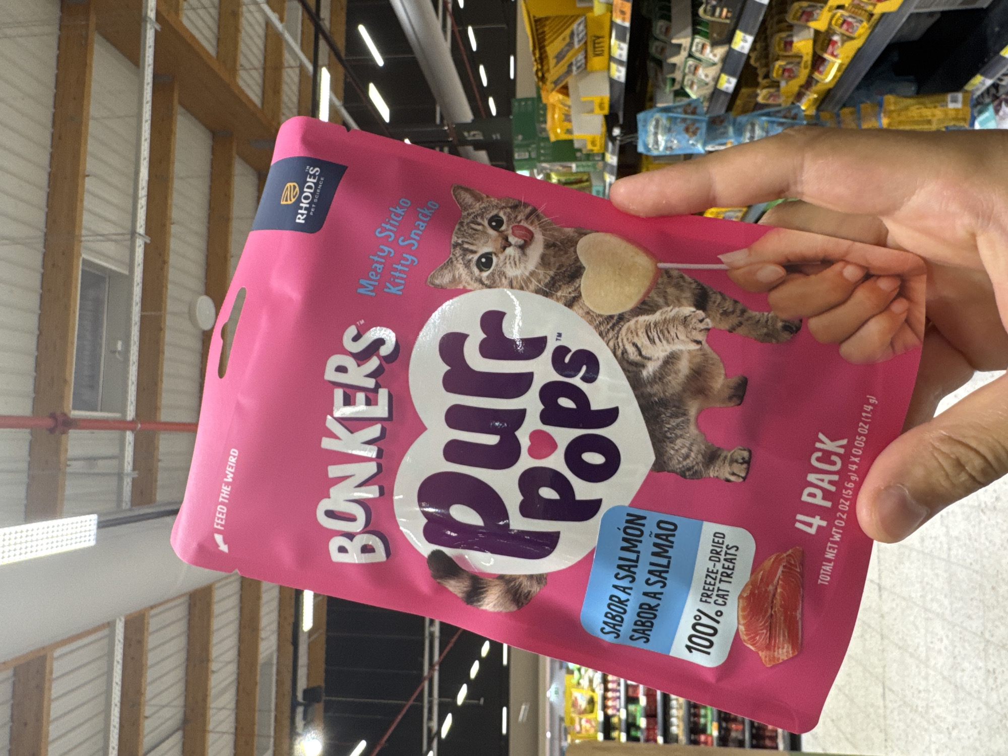 A person is holding a pink package of "Bonkers Purr Pops" cat treats in a store. The package features a tabby cat licking its lips and a heart-shaped treat on a stick. The text on the package indicates the flavor is "Sabor a Salmón / Sabor a Salmão" (Salmon flavor) and that they are "100% Freeze-Dried Cat Treats." The package also states it is a "4 Pack" with a total net weight of 0.2 oz (5.6 g) for 4 x 0.05 oz (1.4 g) treats. The background shows shelves stocked with other pet products in what appears to be a retail environment, possibly a supermarket or pet store. The lighting suggests an indoor setting. There is no specific indication that this scene is located in Castelo Branco, Portugal.