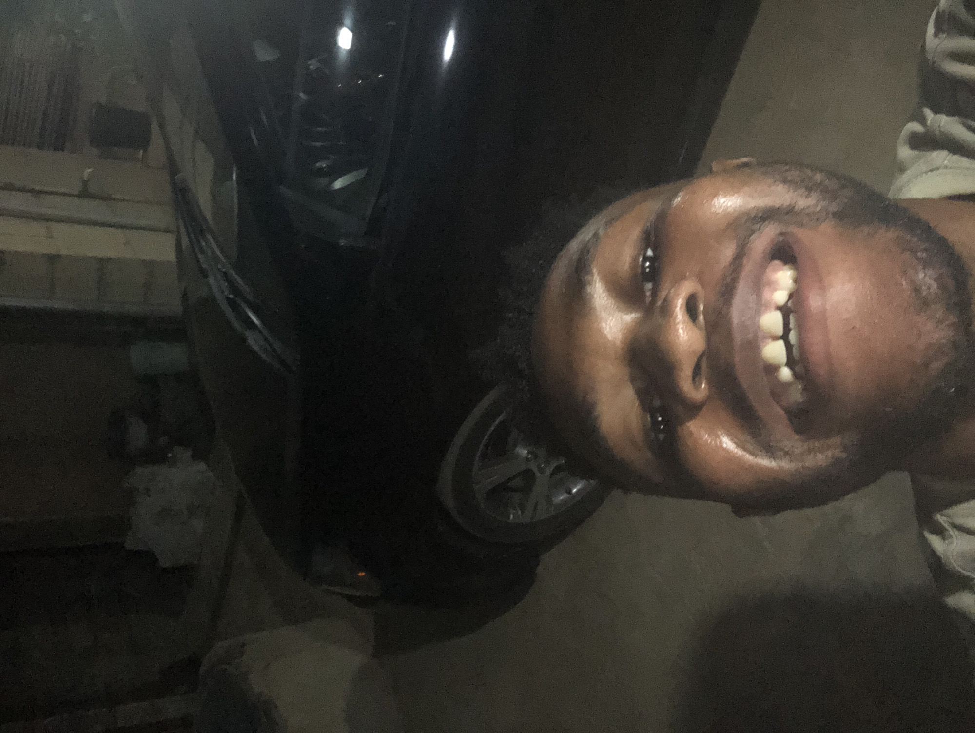 A person with dark skin and a shaved head is smiling. The person is next to a dark-colored car with a silver rim on the wheel. The location appears to be outdoors at night.
