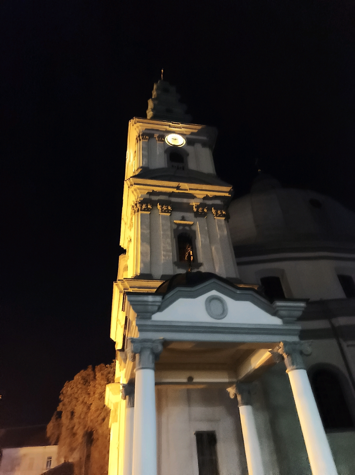The image shows a clock tower of a church or cathedral at night, illuminated by artificial light. The tower is tall and slender, with a pointed spire at the top. A circular clock face is visible on one side of the tower. The building appears to be made of stone or concrete and has architectural details such as columns and decorative elements. To the right of the clock tower, a large dome is visible, suggesting a larger church structure. The sky is dark, indicating it is nighttime. In the foreground, there is an entrance or portico supported by white columns. The overall impression is a majestic religious building at night. This is the Church of the Immaculate Conception of the Blessed Virgin Mary in Ternopil, Ukraine.