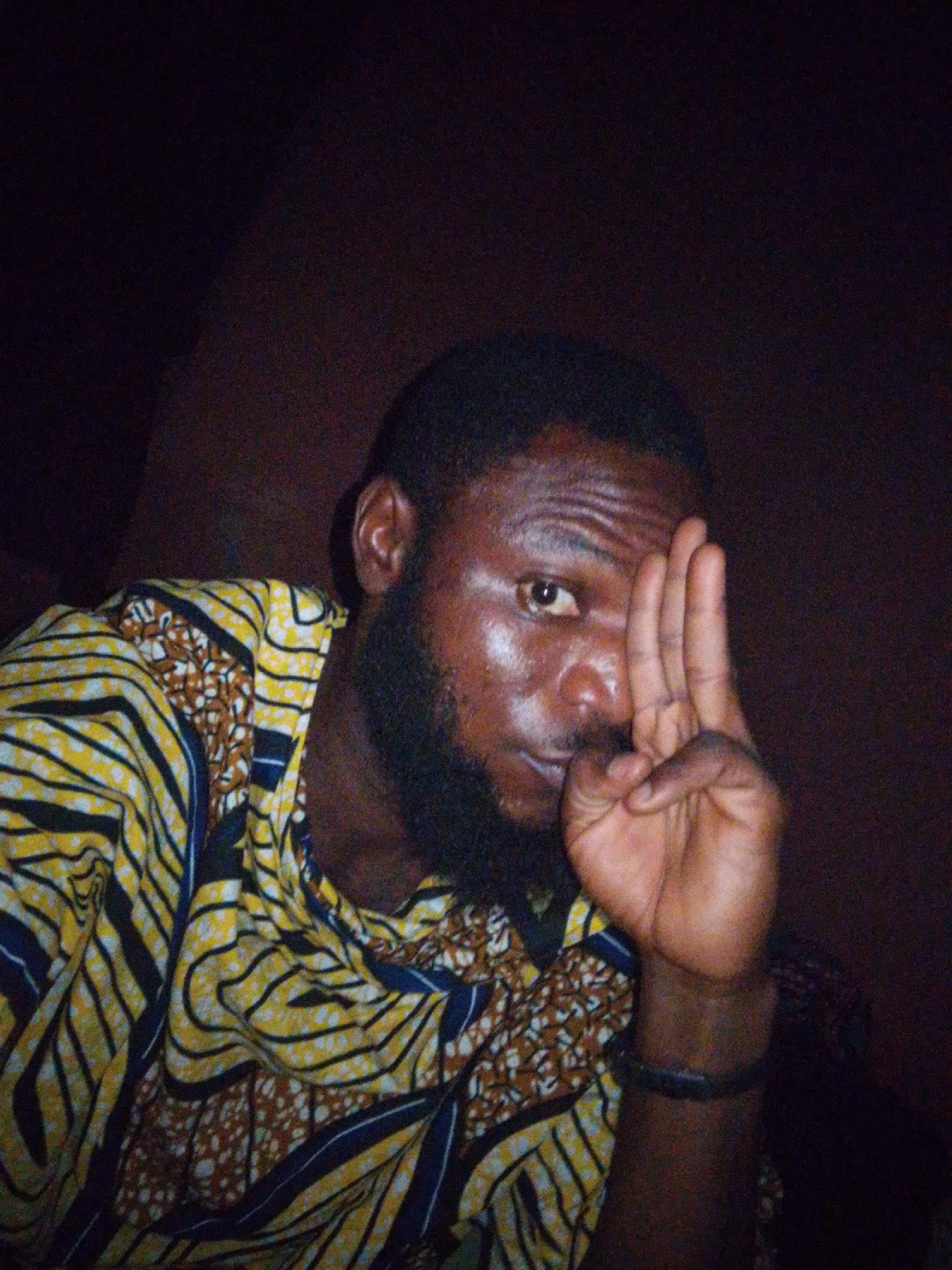 A man is partially obscured, looking at the camera, with three fingers raised on his right hand. He has dark skin, a black beard, and short black hair. He is wearing a patterned shirt with yellow, black, and brown designs. A black wristband is visible on his right wrist. The background appears to be dark and indistinct. The image does not provide enough information to definitively place the subject in Lokoja, Nigeria.