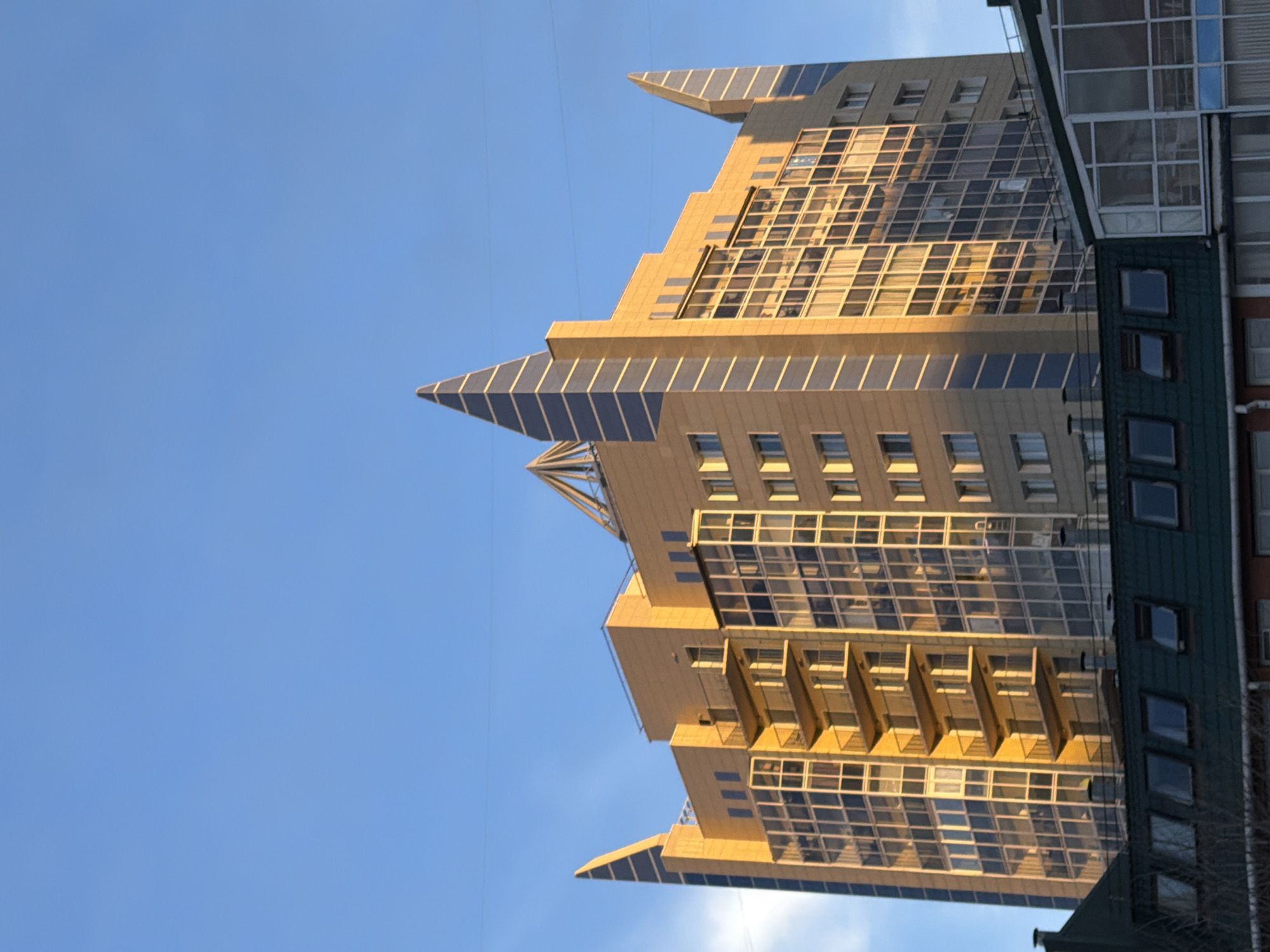 A low-angle, upward-looking shot shows a tall, modern building in Krasnoyarsk, Russia, against a clear blue sky. The building features a unique architectural design with multiple tiers and sharp, triangular protrusions at the top. Its façade is primarily constructed of glass windows arranged in a grid pattern, reflecting the sunlight. The building appears to be adjacent to a more traditional building with smaller, more rectangular windows and a dark-colored exterior. Bare branches of a tree are visible at the bottom corner of the frame. Electrical wires stretch across the upper part of the sky.