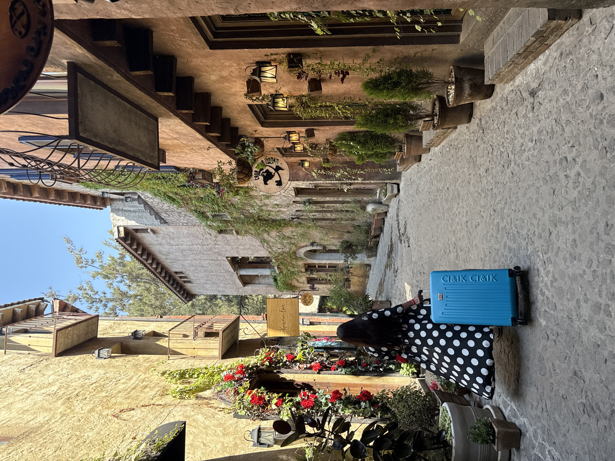 A photograph depicts an alleyway in Domingo Arenas, Mexico. The buildings are constructed of textured stone and stucco in warm hues. The narrow passageway is paved with uneven stone. A person wearing a black-and-white polka dot dress stands with a bright blue suitcase. The buildings are adorned with planters containing red flowers and various green foliage. Lighting fixtures are affixed to the buildings. Signage is visible, including a round sign reading "CASA". There is a wooden sign reading "Karina Love". The sky is blue and partly visible.