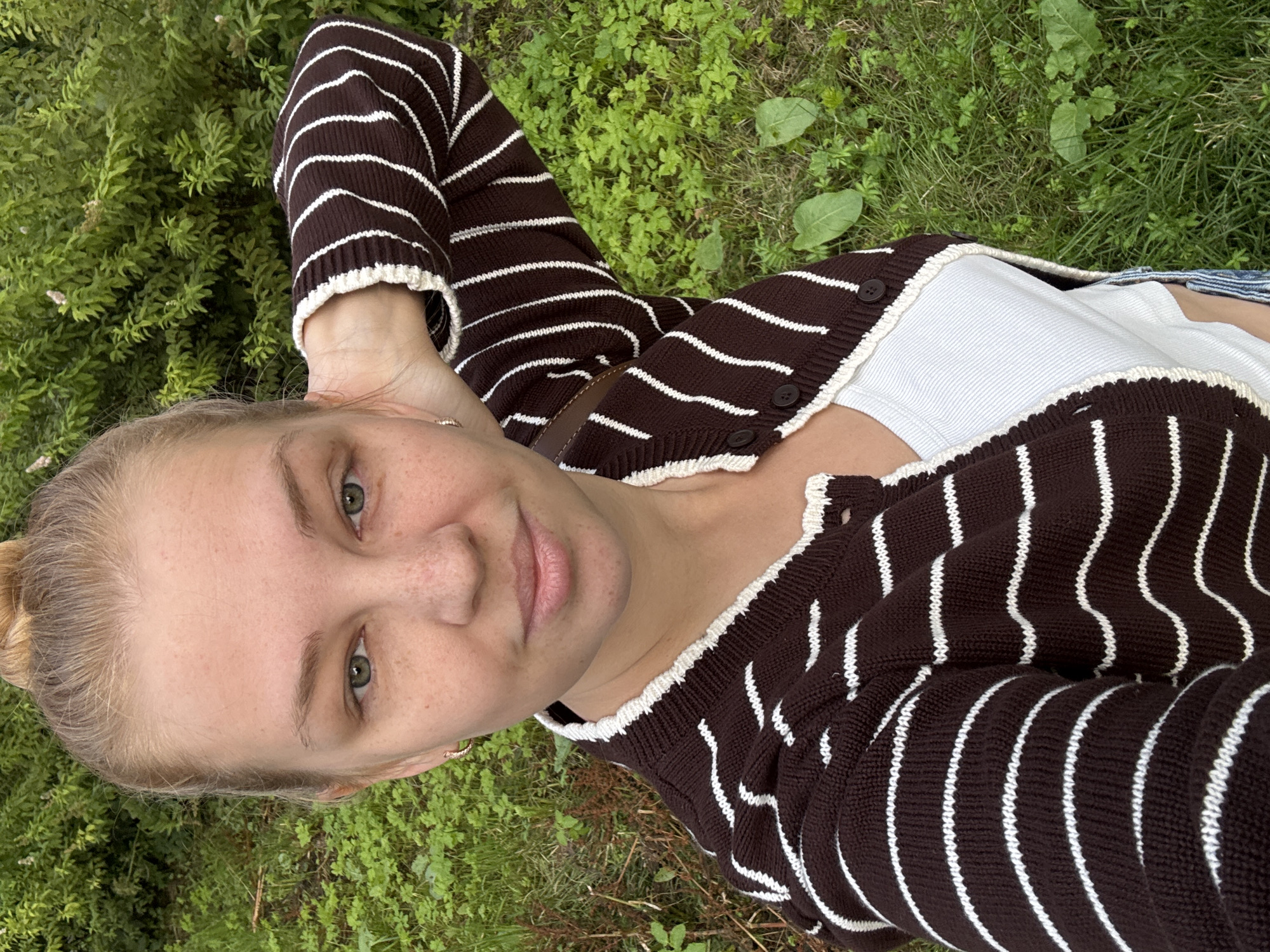 A young Caucasian woman with blonde hair in a bun is standing outdoors in a grassy area with green bushes in the background. She is wearing a brown and white striped cardigan over a white tank top and appears to be taking a selfie. Her left arm is raised and bent, with her hand behind her head. The lighting suggests it is daytime. There are no discernible landmarks that would indicate a specific location within Rusanovo, Russia.