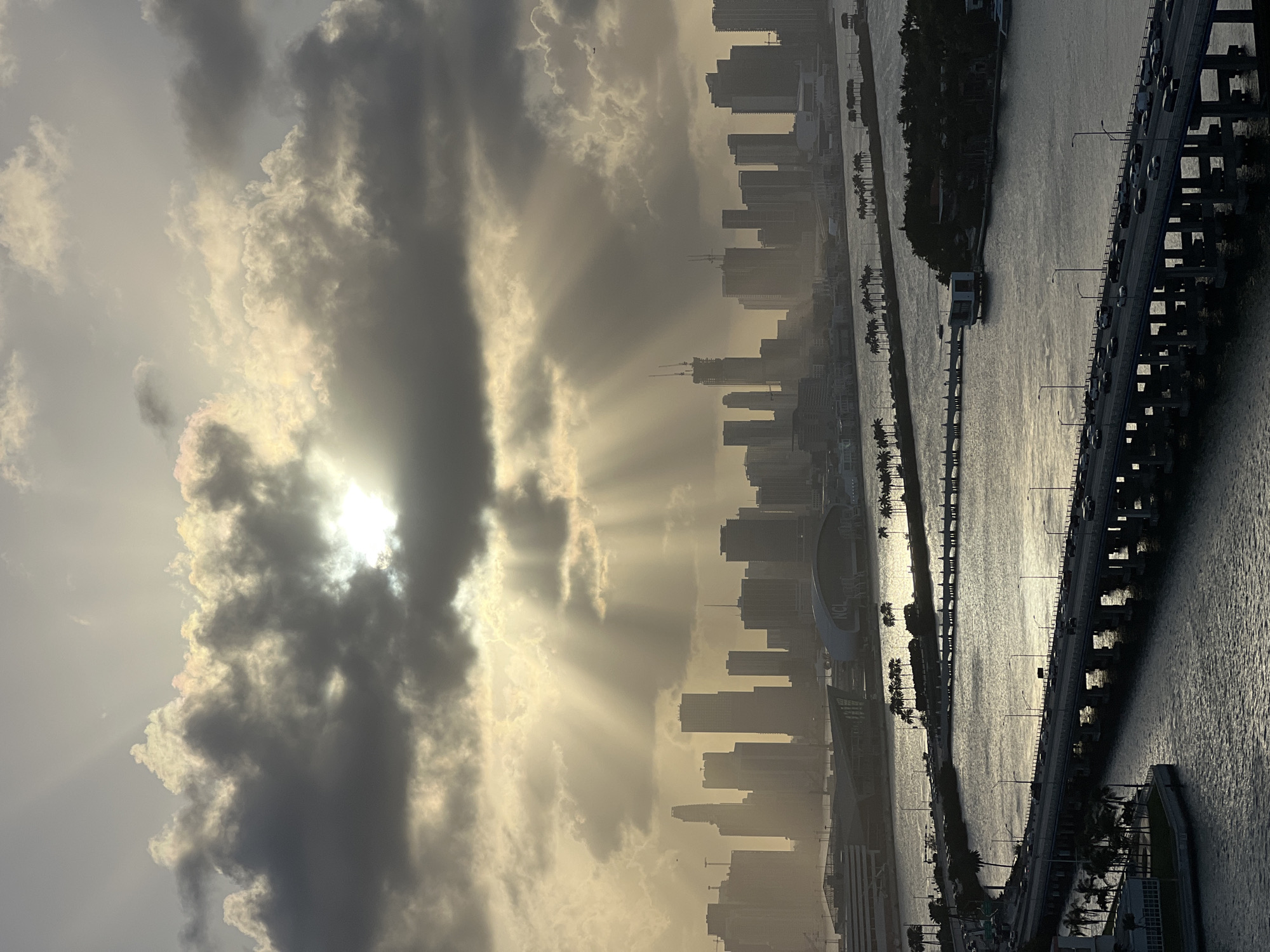 The image captures a sunrise over Miami Beach, Florida, United States. A dramatic sky with heavy clouds obscures the sun, but its rays break through, casting light beams over the city skyline. Buildings are silhouetted against the sunlit sky, lining the shore. A bridge extends across the water, with cars visible. The water reflects the sky's light, creating a shimmering effect. Palm trees line the waterfront, adding a tropical touch to the urban landscape.