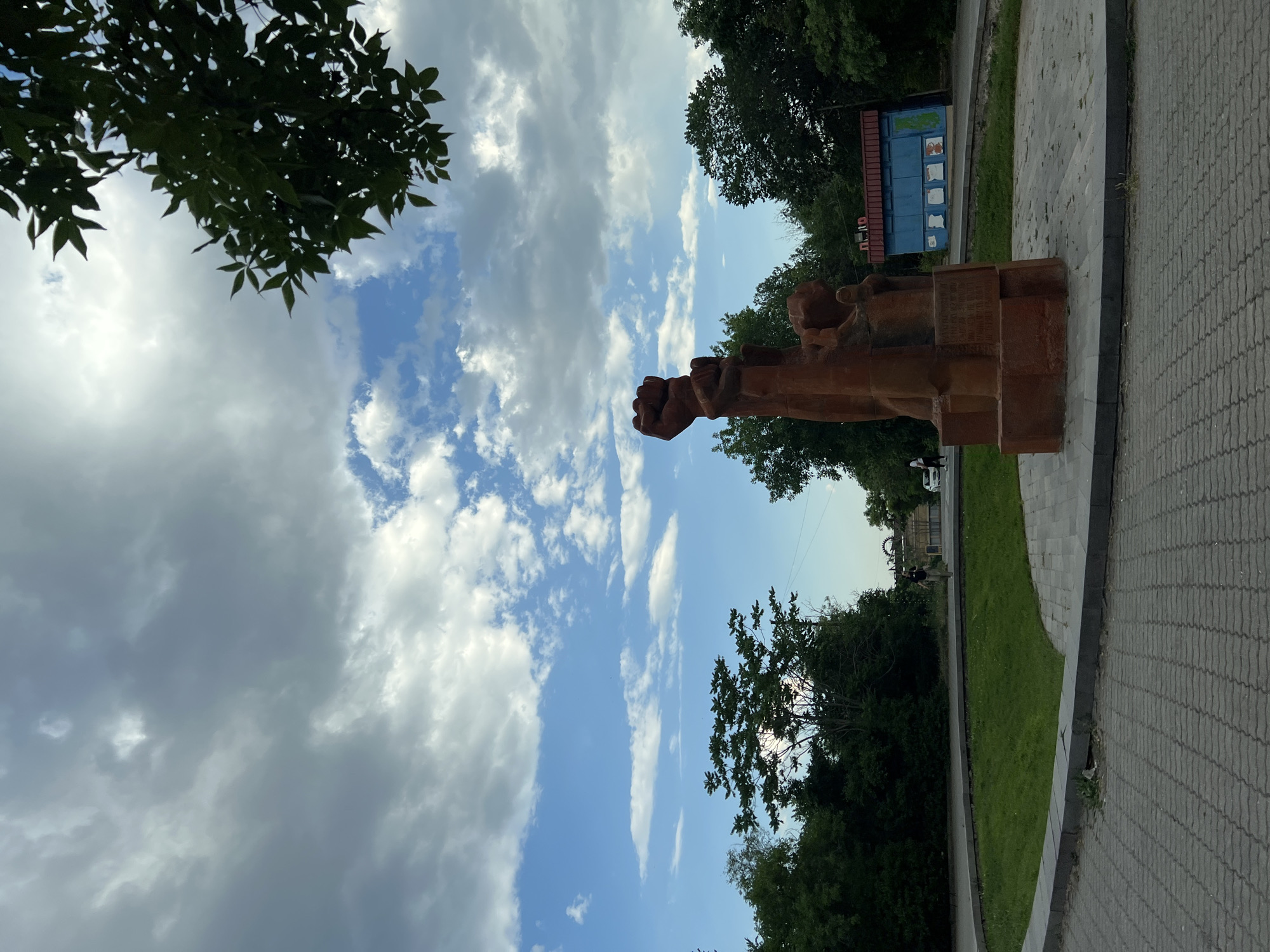 This image, taken in Yerevan, Armenia, captures a statue of a clenched fist made of a reddish-brown material. The statue appears to be made of stacked blocks, with the fist prominently raised. It is situated outdoors, with a paved walkway and a grassy area in the foreground. Lush green trees and a bright blue sky filled with dramatic, fluffy clouds dominate the background, suggesting it's daytime. Sunlight filters through the clouds, creating a well-lit scene.
In the distance, a small blue structure with red trim is visible, possibly a kiosk or small building, with some text or images on its side. The overall atmosphere seems peaceful, with the focus on the symbolic statue against the backdrop of nature. The composition, with the sky taking up a large portion of the frame, emphasizes the grandeur of the clouds and the sky. No specific activities or events are clearly depicted, but the presence of the statue suggests a public space, possibly a park or memorial area. The time of day appears to be afternoon or late morning, given the brightness and the cloud formations.