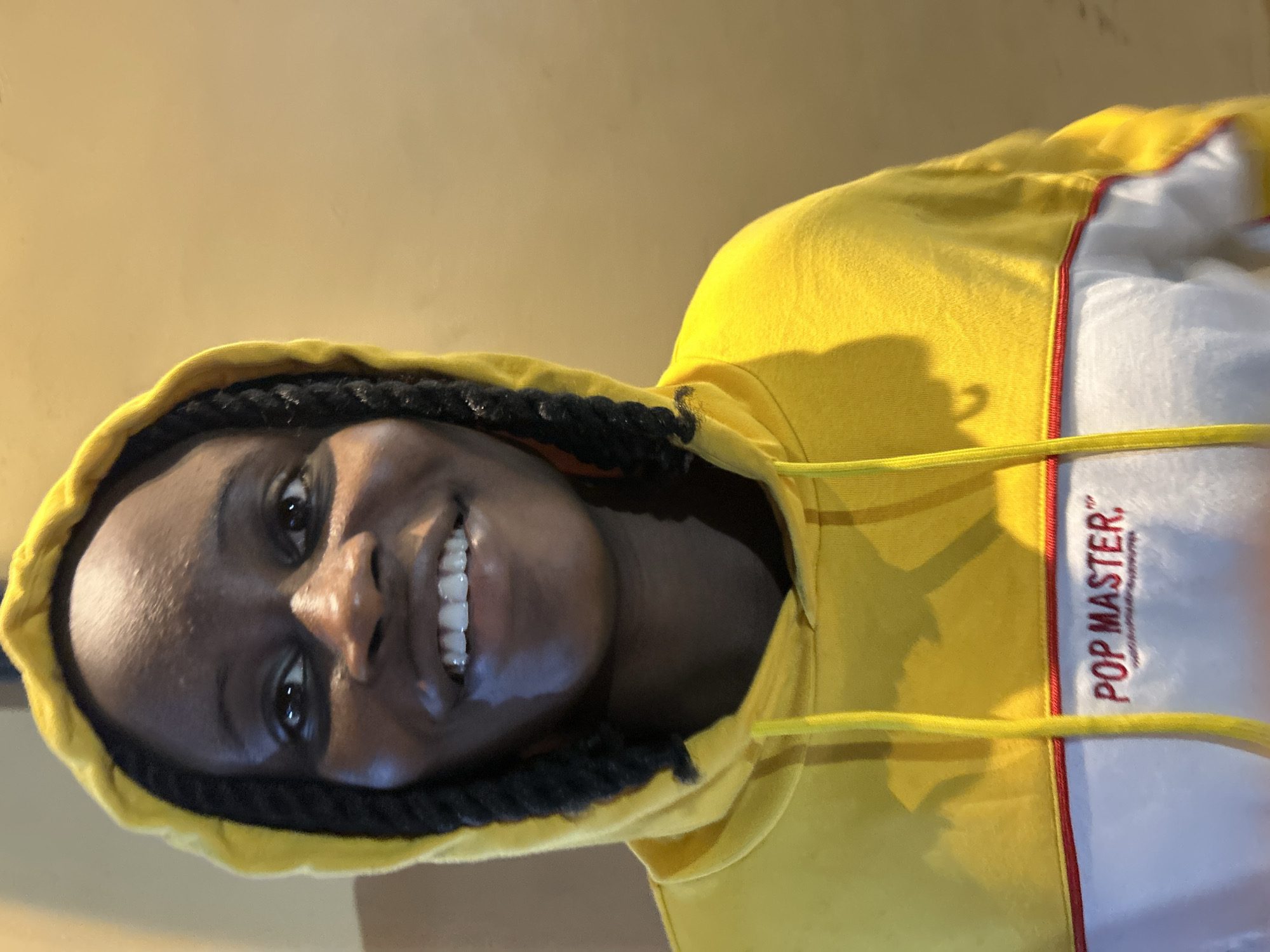 A head and shoulders portrait shows a young Black woman wearing a yellow hooded sweatshirt with white and red accents and the text "POP MASTER" embroidered in red on the white section. She has dark braided hair and is smiling broadly, showing her teeth. The background is a plain, light brown wall. The image was captured in Ihiagwa, Nigeria.