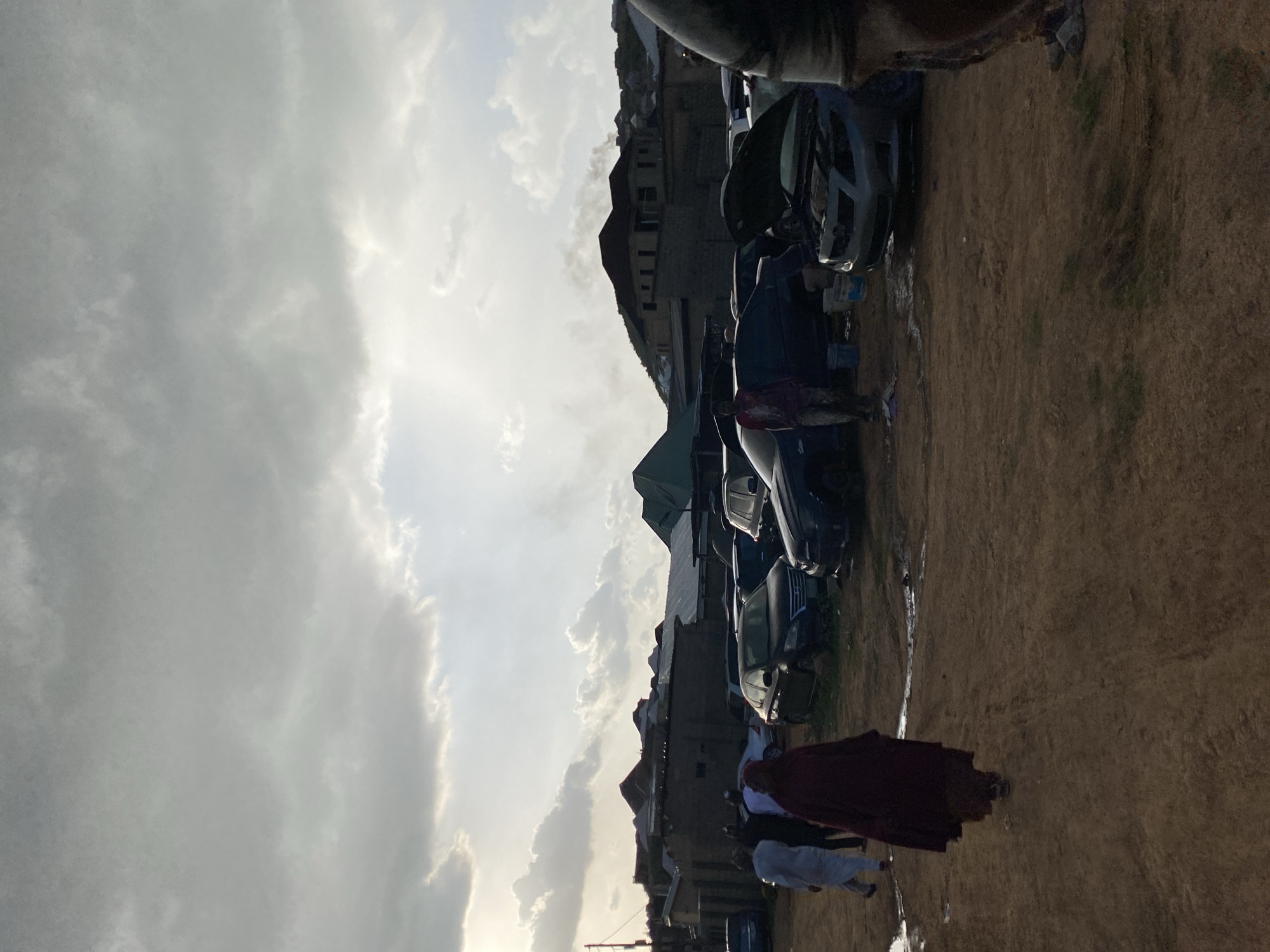 The image depicts a scene in Jos, Nigeria, with several cars parked on a dirt lot. The sky is filled with dramatic, overcast clouds, suggesting it might be a cloudy or stormy day. The lighting indicates it is likely late afternoon or early evening.
In the foreground, several people are visible. One person is wearing a maroon garment, possibly a dress or robe, and is walking away from the camera. Another person behind them is wearing a white outfit. The people appear to be casually walking or gathering.
Behind the people and cars, there are buildings with pitched roofs, some appearing to be made of stone or concrete. The overall setting suggests an outdoor urban or semi-urban environment, possibly a marketplace or a residential area with open space for parking.
The image is oriented vertically, with the sky occupying the upper portion and the ground with cars and people in the lower portion. The ground is a dusty, dry earth color. There are no visible text elements in the image. The mood appears neutral, with no strong emotional cues evident.