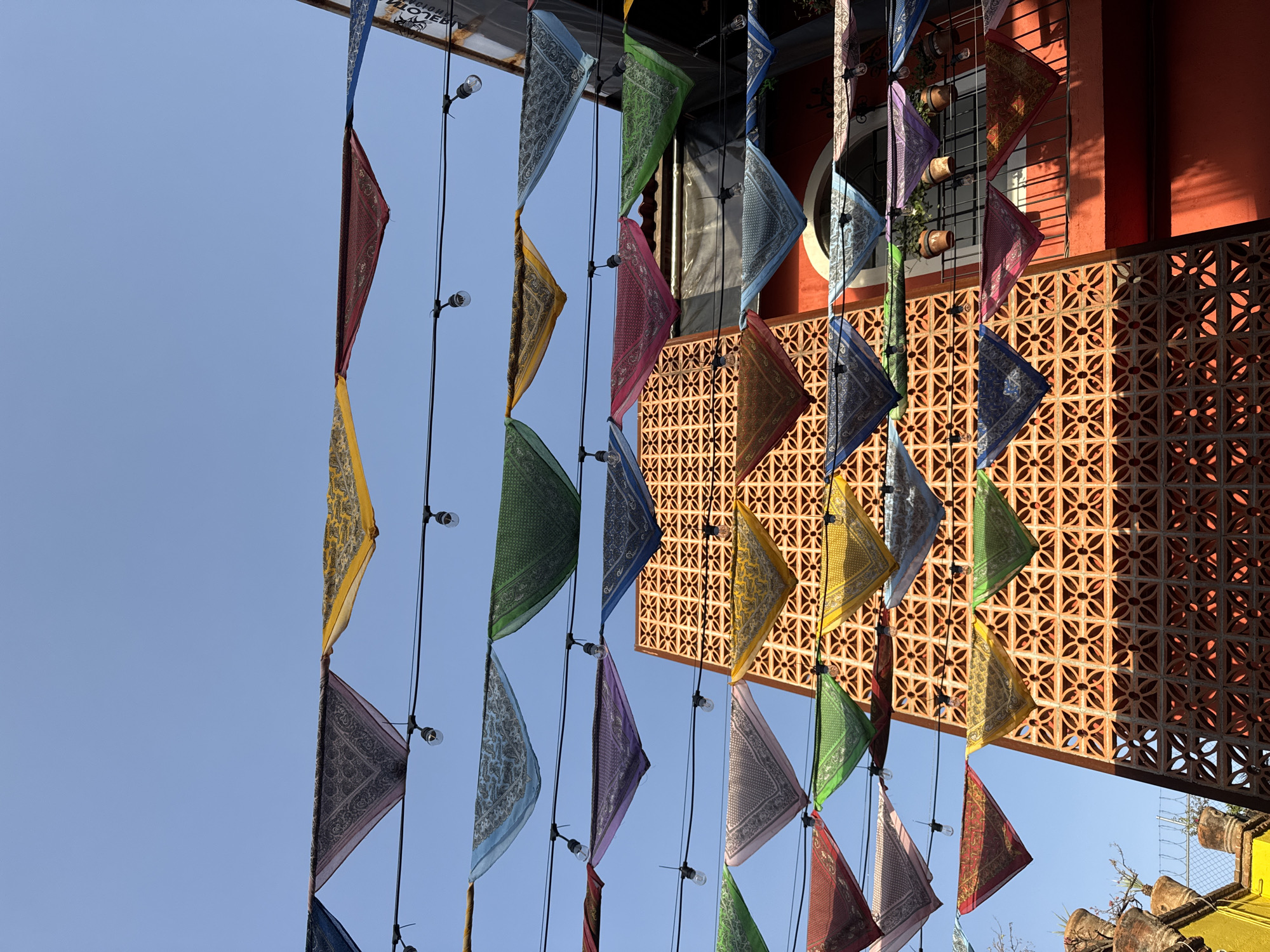 A street scene in Puebla, Mexico, is captured from a low angle, featuring a row of colorful triangular flags or bandanas hanging from a string of lights against a clear blue sky. The flags create a vibrant display of patterns and colors. In the background is a red building with a lattice brick or tile accent wall, suggesting architectural detail typical of the region. Plants in terracotta pots are visible atop the wall. The overall impression is a sunny, festive, and culturally rich urban environment.