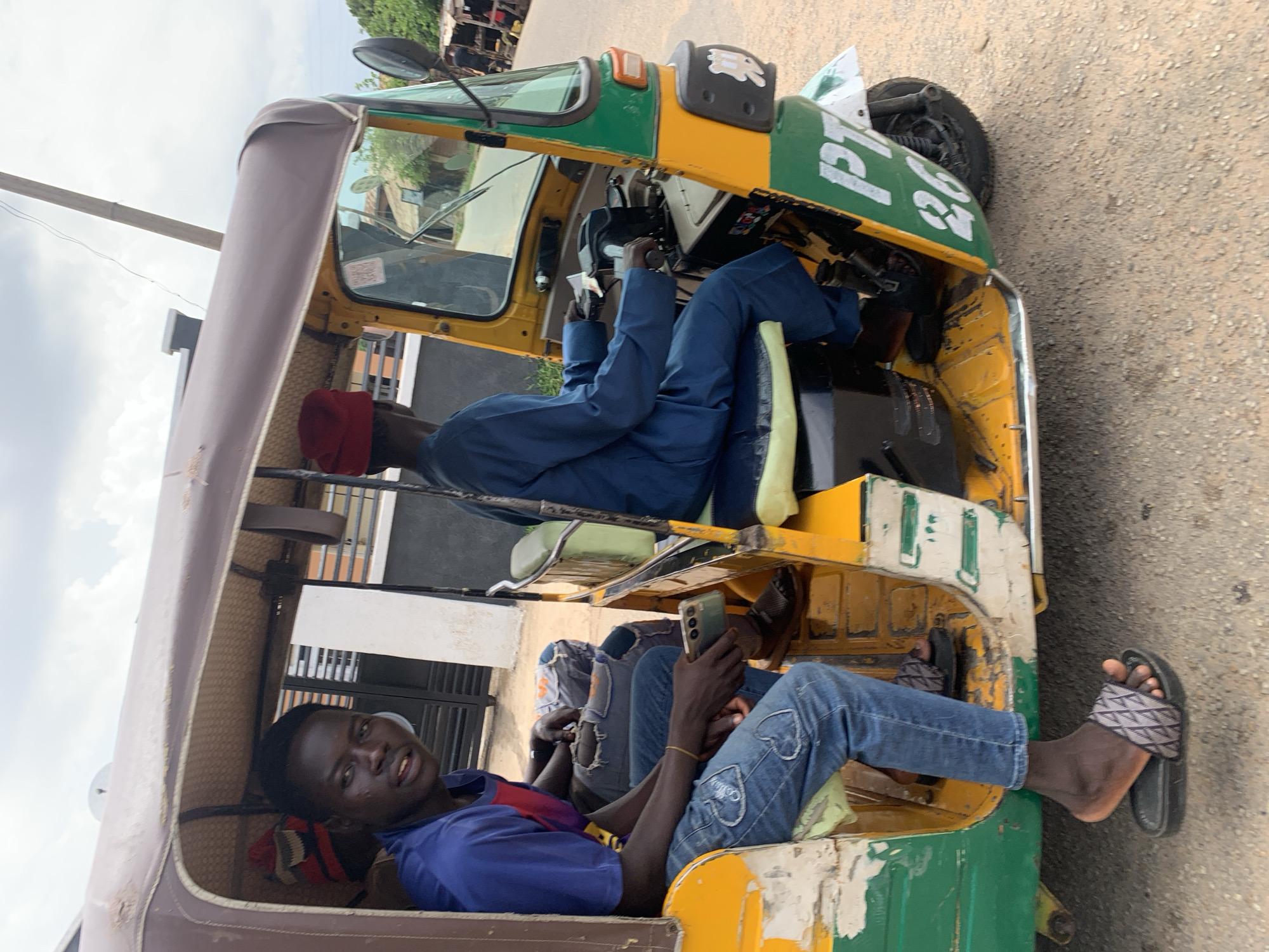 This rotated image displays a three-wheeled auto-rickshaw, commonly known as a Keke NAPEP, on a paved road, likely in Jos, Nigeria. The vehicle features a distinctive green and yellow paint scheme with a brown canvas canopy, showing signs of significant wear and chipping.
Inside the Keke, three individuals are present. The driver, dressed in a blue suit and a red cap, is seated at the front, his hands on the handlebars. Behind the driver, a male passenger is positioned comfortably, leaning back. He wears a blue short-sleeved shirt and blue jeans adorned with white embroidered heart shapes. He holds a smartphone in his hands and gazes towards the camera with a relaxed, slight smile. His left foot, wearing a black slide sandal with a patterned strap, extends out of the vehicle. A third passenger is partially visible behind the first, also wearing jeans.
The setting suggests a daytime scene under an overcast sky. In the background, glimpses of buildings and green foliage can be seen. Visible text on the front section of the Keke, painted in white, reads "PT 26". The overall impression is a candid moment of daily life and transportation in the region.