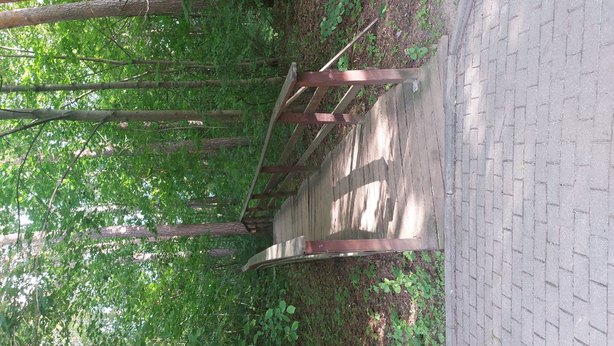 The image depicts a wooden bridge with brown railings in a wooded area, likely a park or forest. The bridge connects to a paved surface made of rectangular stones. The environment consists of trees with green foliage and ground cover. The location is likely Khimki, Russia. There are no people visible in the image, and no specific actions are evident. The scene is static and focused on the bridge and its immediate surroundings.