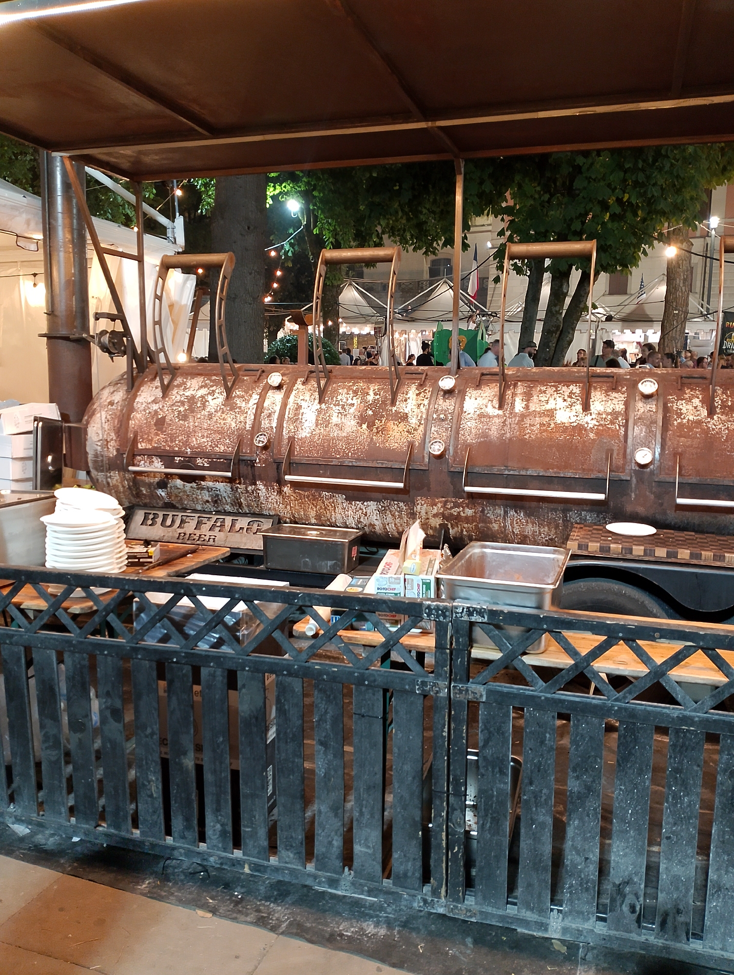 The image shows a large, rustic smoker, likely a barbecue pit, situated outdoors. The smoker is made of metal and appears to be quite old, with visible signs of rust and weathering. It has several doors or compartments along its length, each with a handle. A stack of white plates is visible to the left of the smoker, along with a wooden sign that reads "BUFFALO BEER." A metal grate fence runs across the foreground, separating the viewer from the smoker and the area behind it, where tables and some people are faintly visible in the background. String lights are present in the background, suggesting an evening or nighttime setting, possibly at an outdoor event or food stall. The overall impression is one of an informal, perhaps food-focused, gathering. The presence of "Buffalo Beer" on the sign might indicate a connection to American-style barbecue, and the term "beer" suggests that alcohol is being served. The location is specified as Rieti, Italy.
