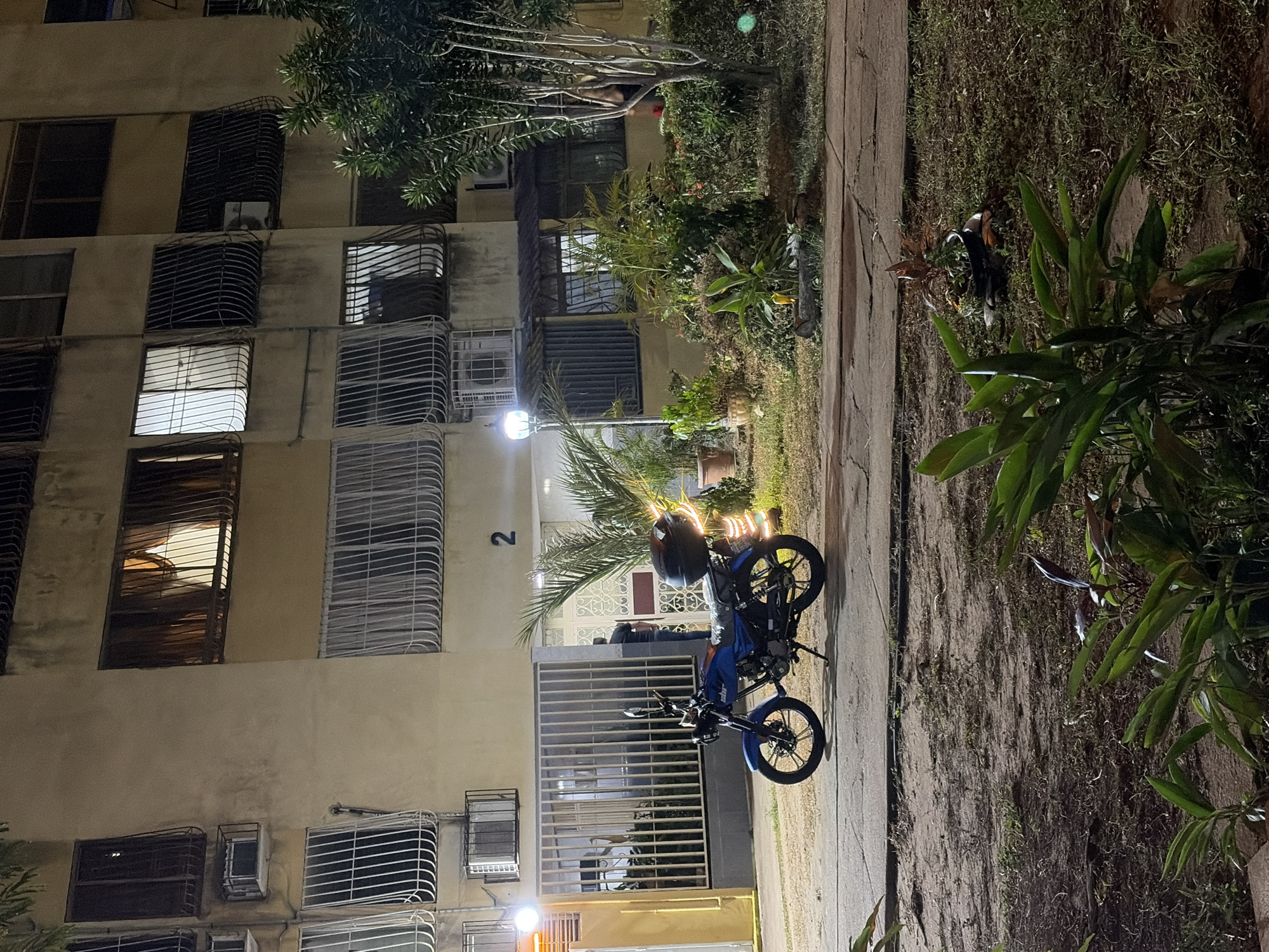 Captured at night in Ciudad Guayana, Venezuela, the image showcases a multi-story apartment building. A blue motorcycle parked in front of the building is a focal point. The building's facade exhibits multiple windows, many with security bars and air conditioning units. Adjacent to the building, a sidewalk runs alongside a grassy area with various plants and trees. The lighting suggests a mix of streetlights and illumination from within the building itself. People are visible at the entrance to the building.